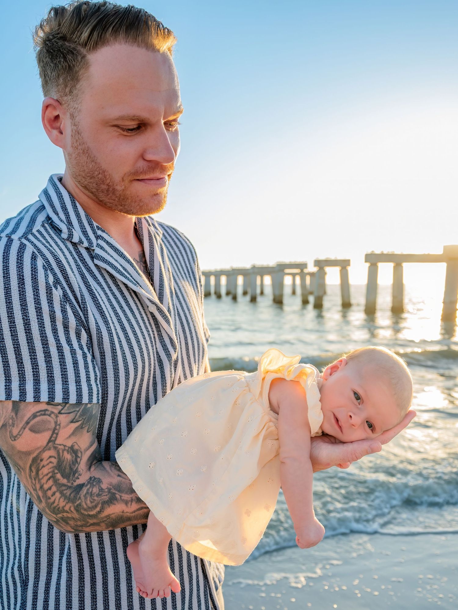 Tattooed father holding newborn baby at sunset beach with pier in background.