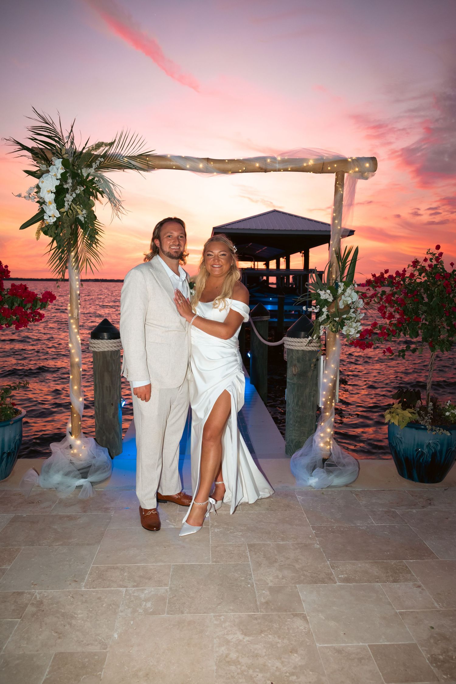 Newlyweds pose under a floral arch with string lights at a stunning waterfront sunset wedding venue.