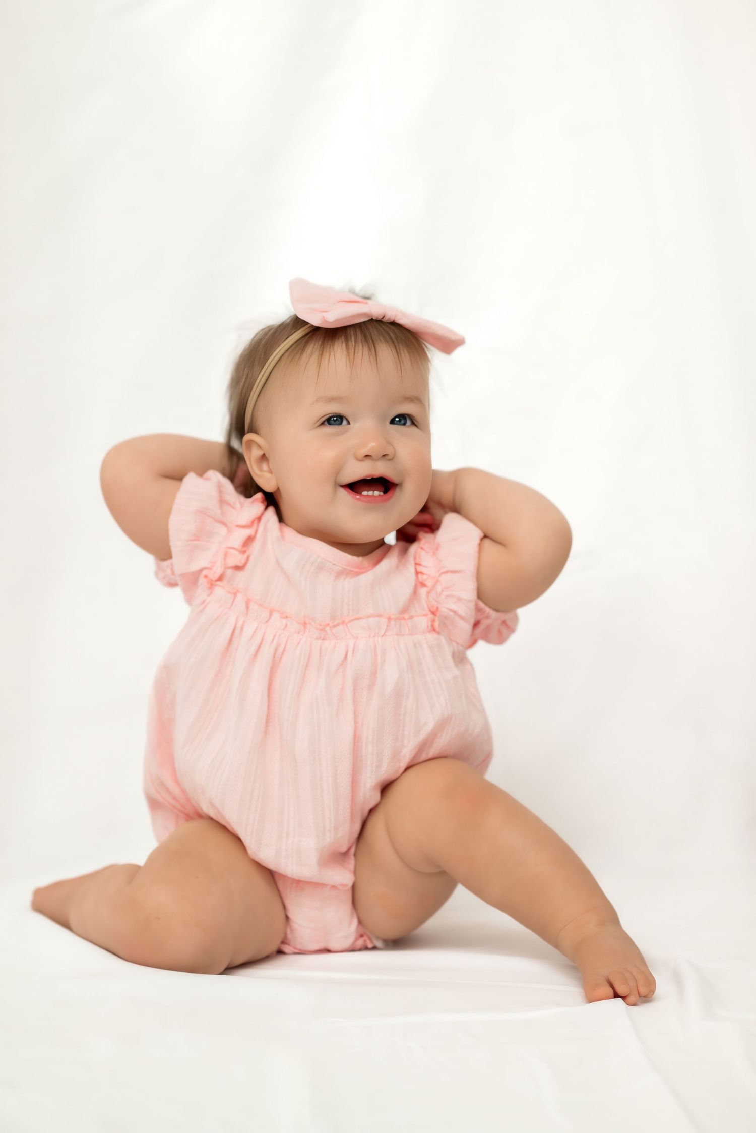 Smiling baby girl in pink ruffle romper and bow headband sitting on white background.