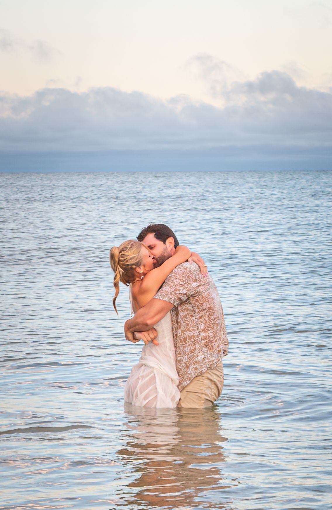 Couple embracing and kissing while standing in shallow ocean water at sunset, romantic beach moment.