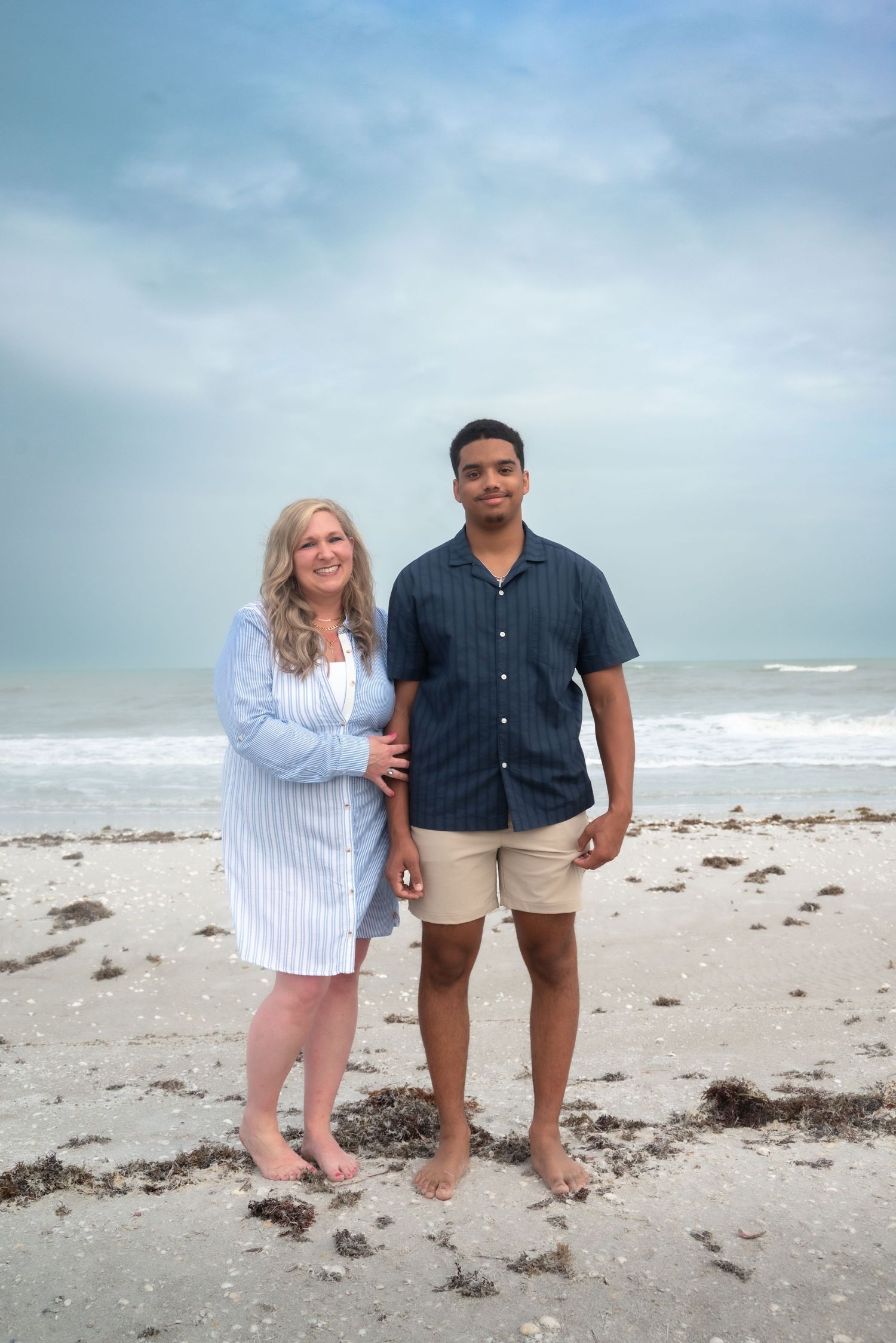 A couple stands barefoot on a sandy beach with ocean waves behind them on a cloudy day.