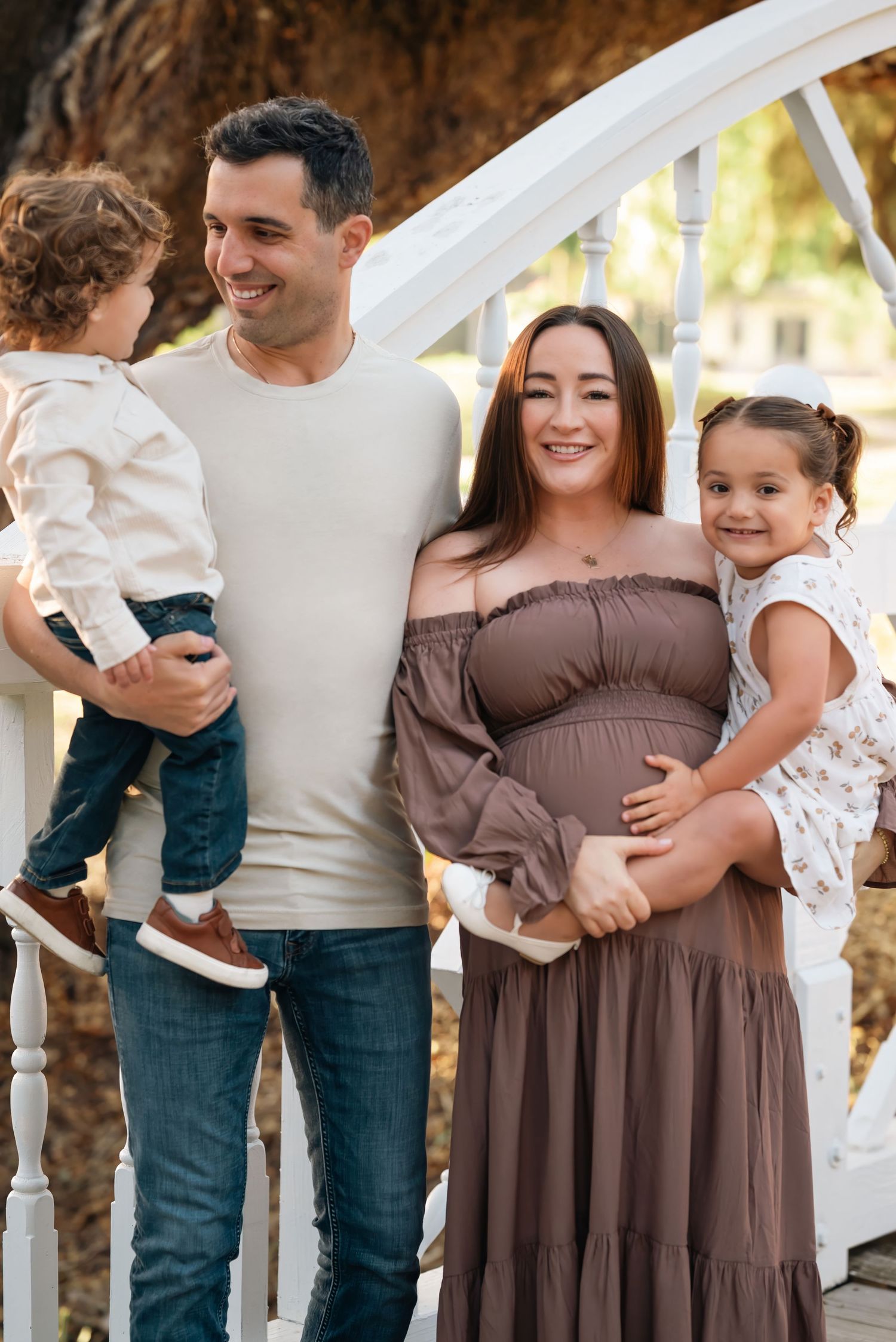 Smiling pregnant woman in brown dress poses with husband and two young children on a white bridge outdoors.