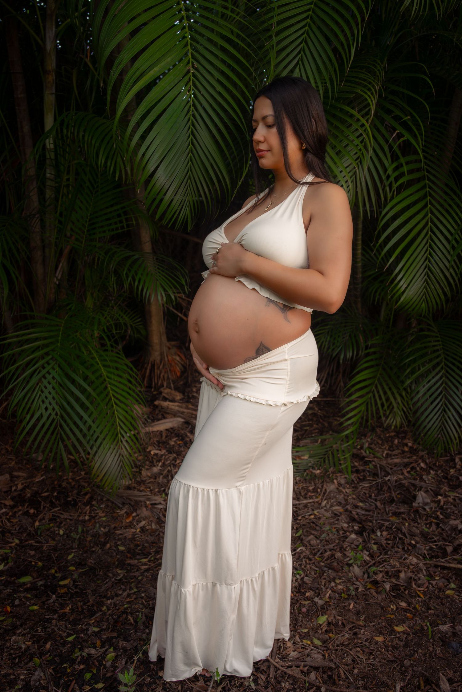 A pregnant woman in a white maternity dress cradles her baby bump surrounded by lush tropical palm trees.
