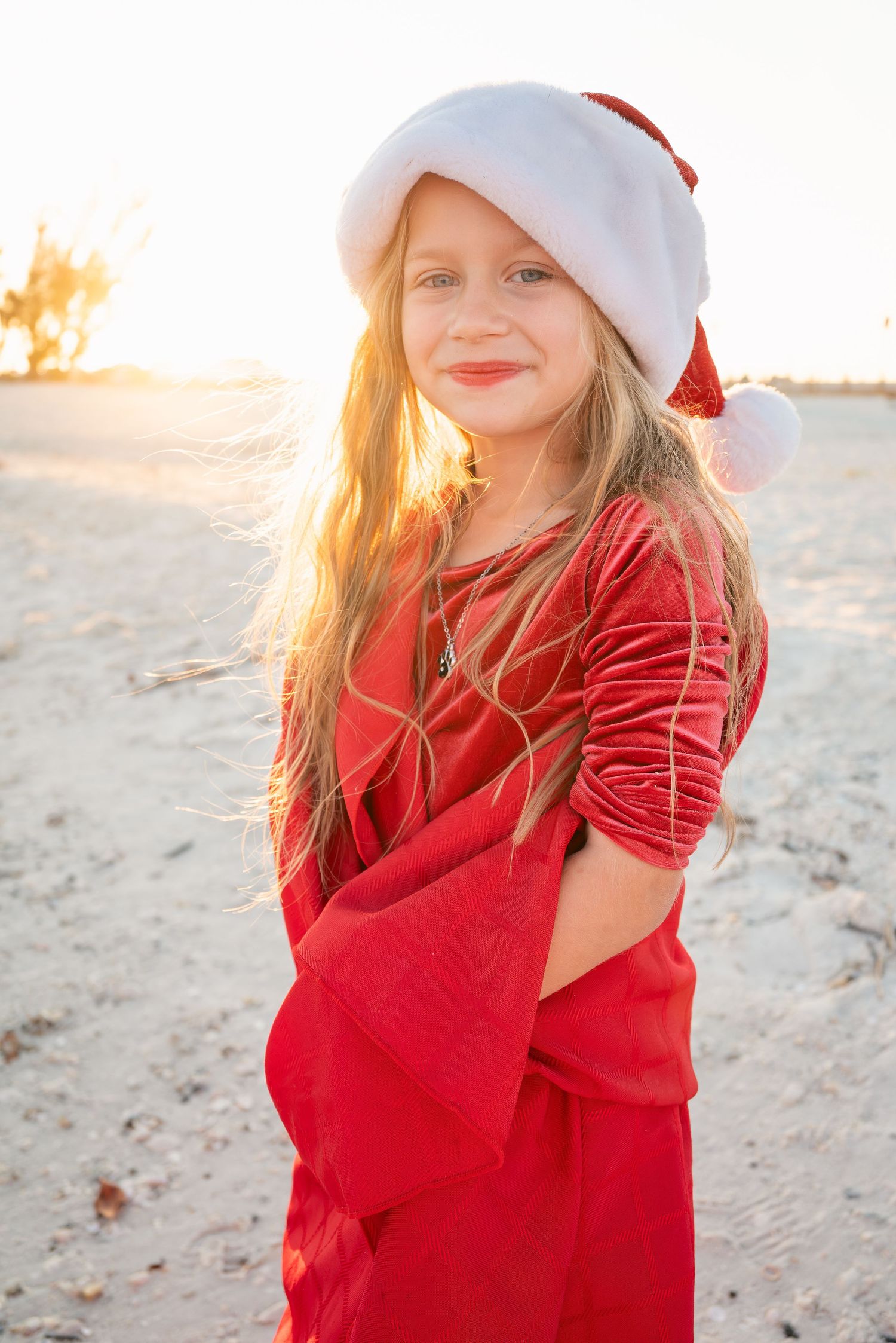 Young girl in red velvet dress and Santa hat smiling on a sunlit beach.