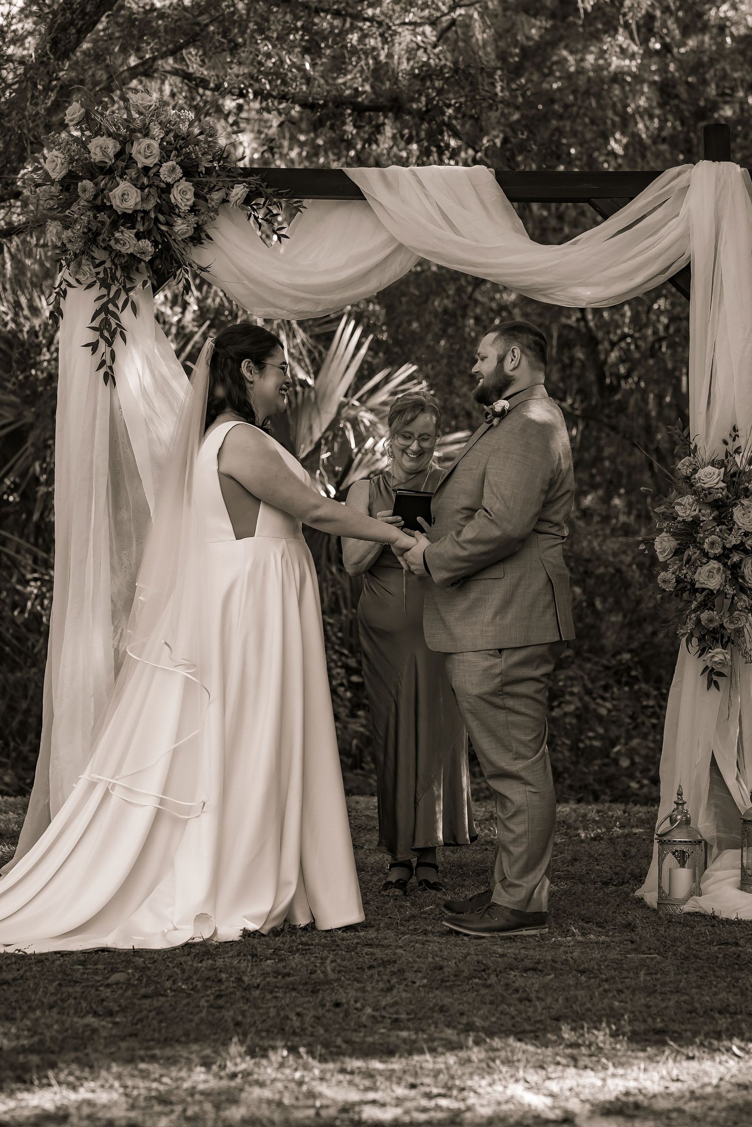 Bride and groom exchange vows under a floral arch with officiant in sepia tone.