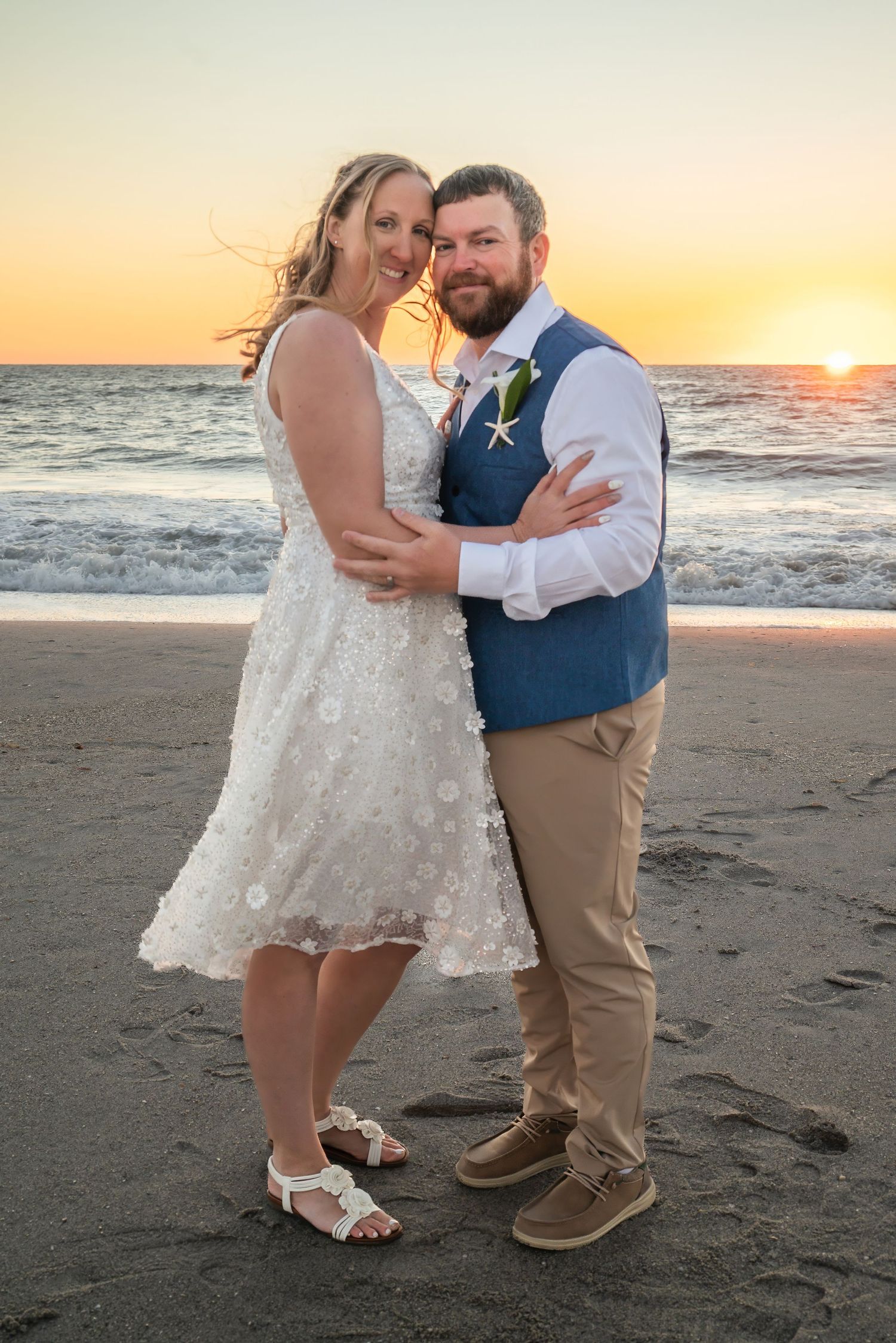 Bride and groom embracing on beach at golden sunset with ocean waves behind them.