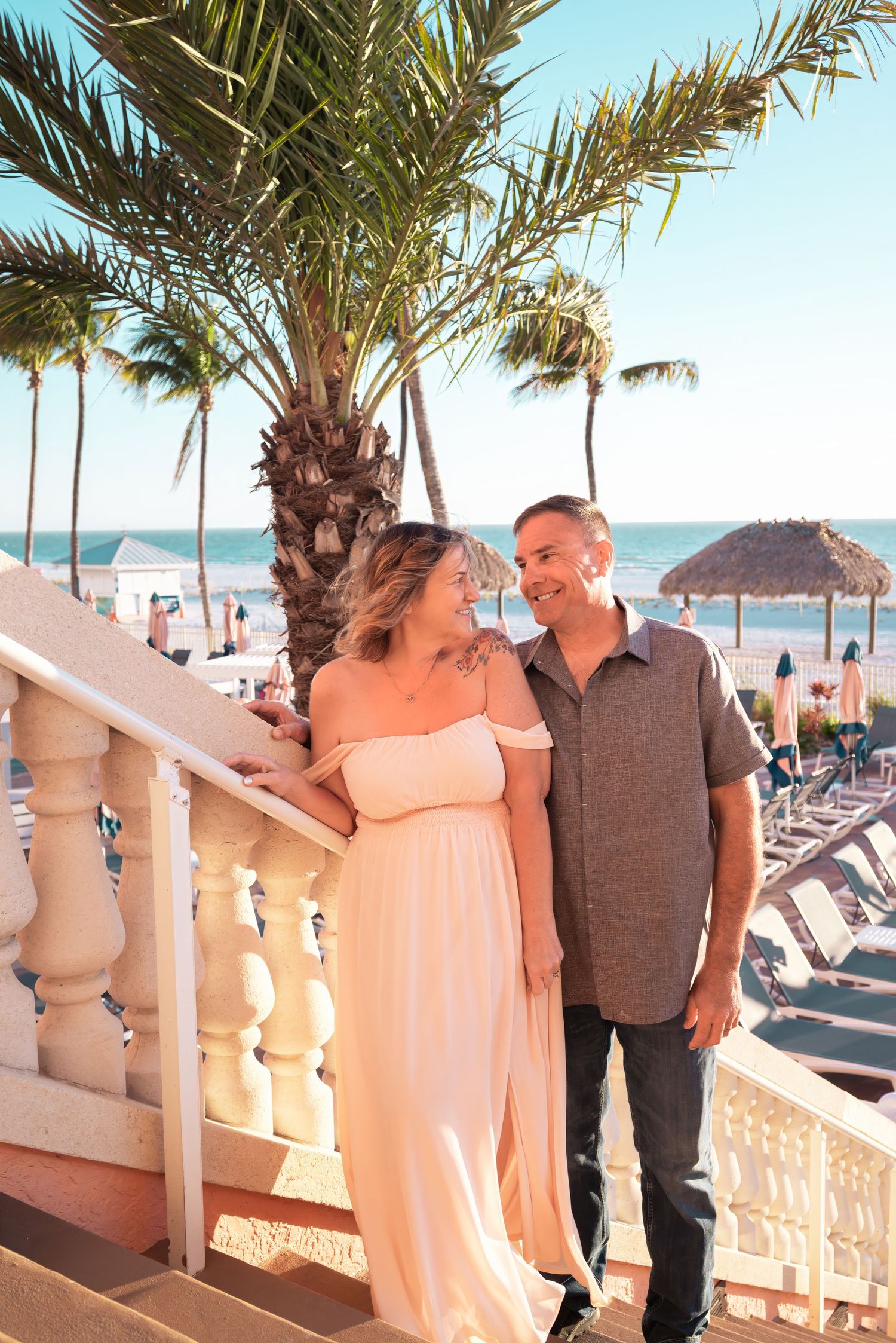 Couple smiling together on a tropical resort balcony with palm trees and ocean in the background.