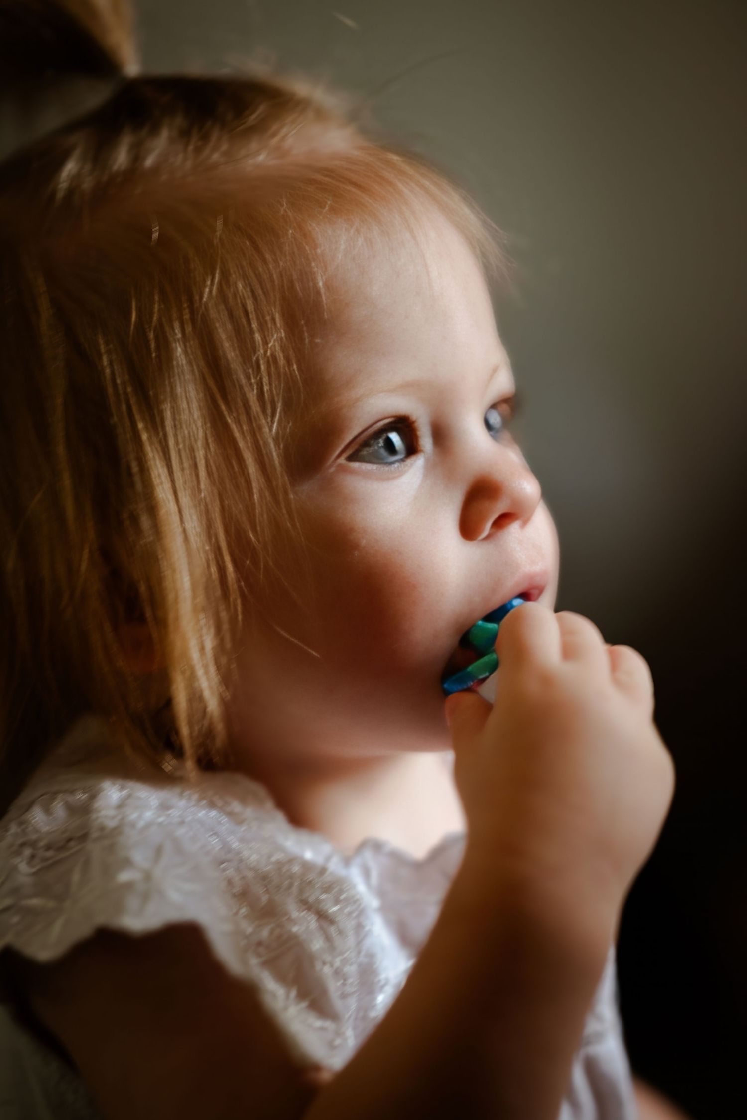A young toddler girl with blonde hair in a white dress holds a colorful toothbrush to her mouth.