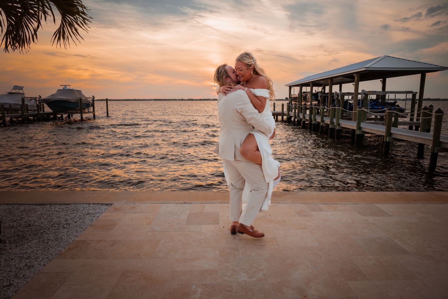 Groom lifts bride at sunset by a waterfront dock, romantic outdoor wedding photo at golden hour.