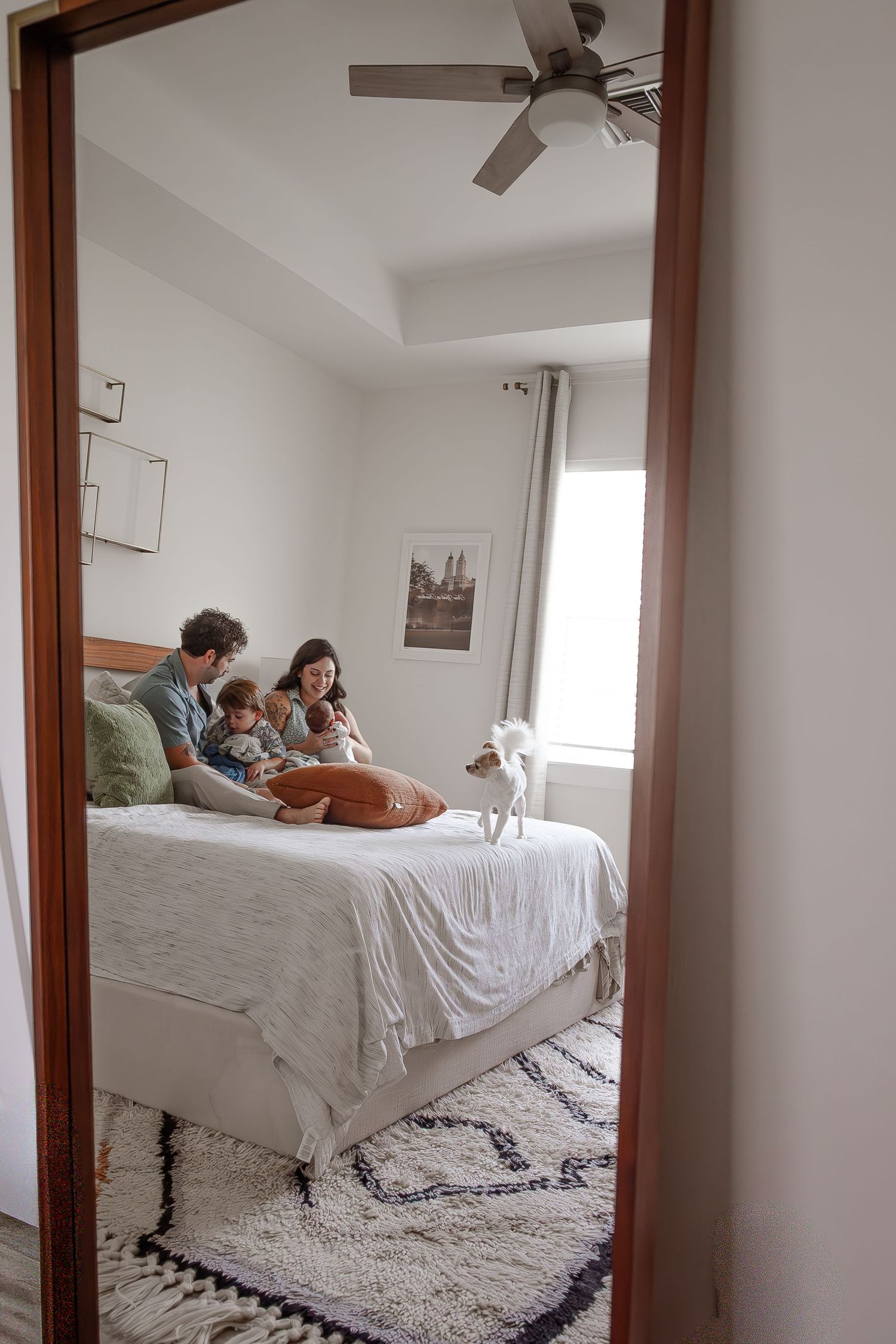 Family of four with small dog relaxing together on white bed viewed through bedroom doorway.