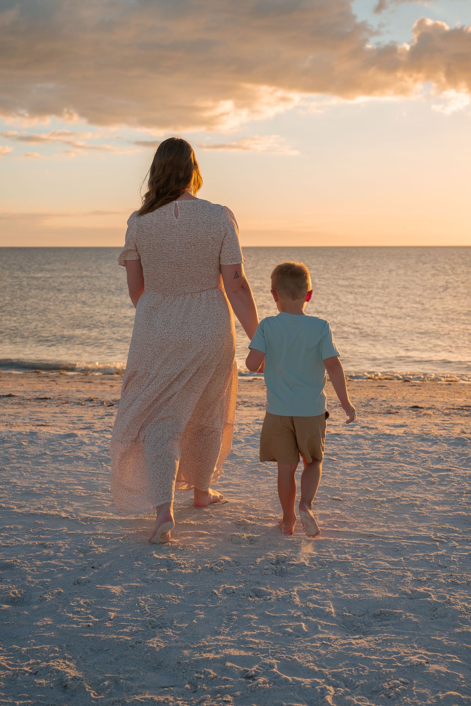 Mother and young son holding hands walking toward the ocean at golden sunset on beach.