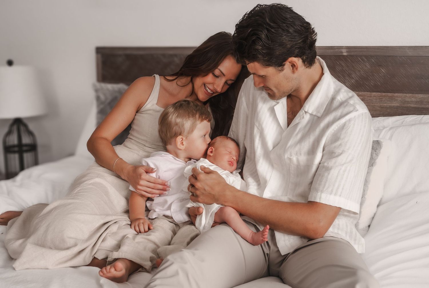 Happy family of four sitting on bed as toddler meets their new baby sibling.
