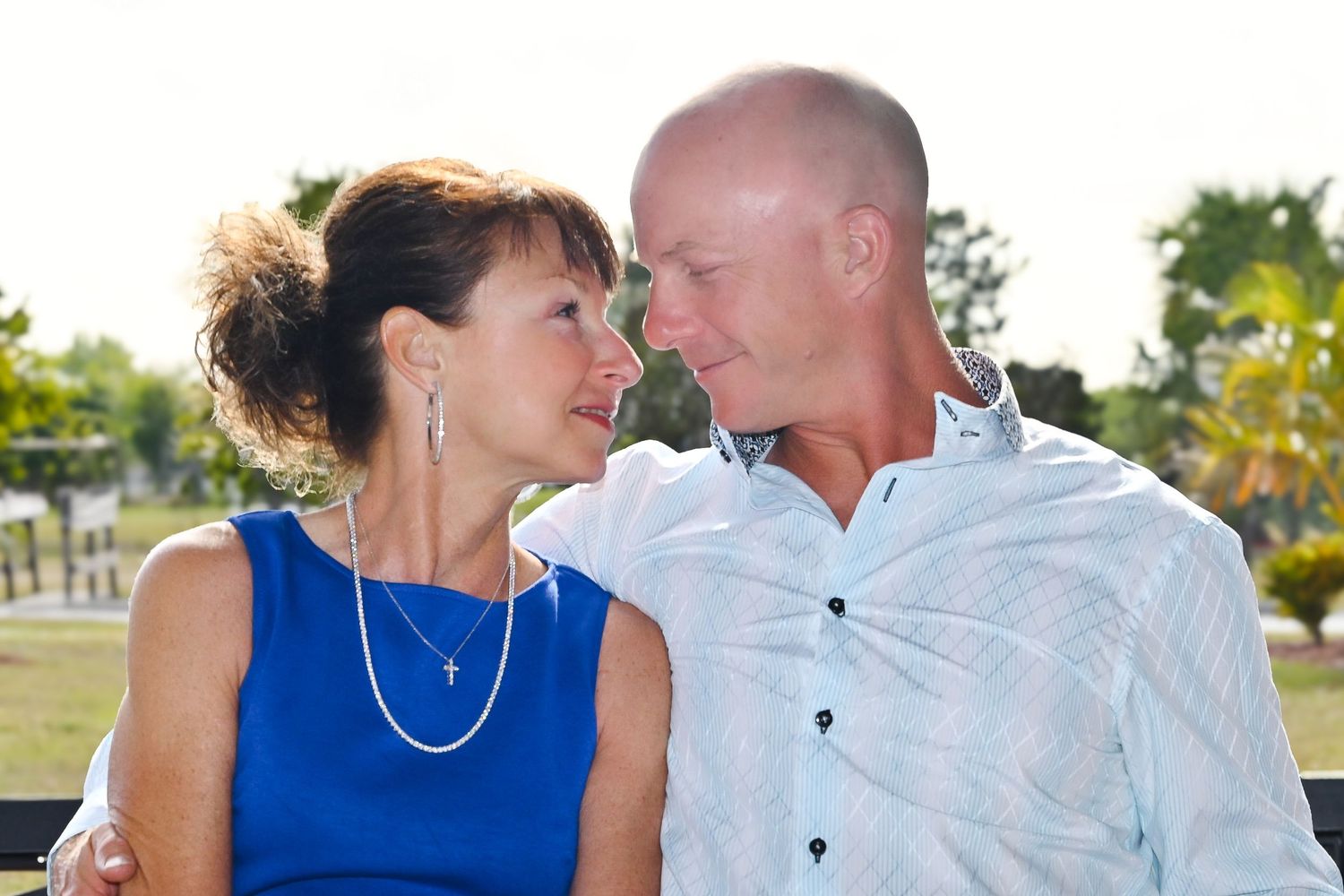 Mature couple sitting close together outdoors, smiling at each other in warm sunlight.