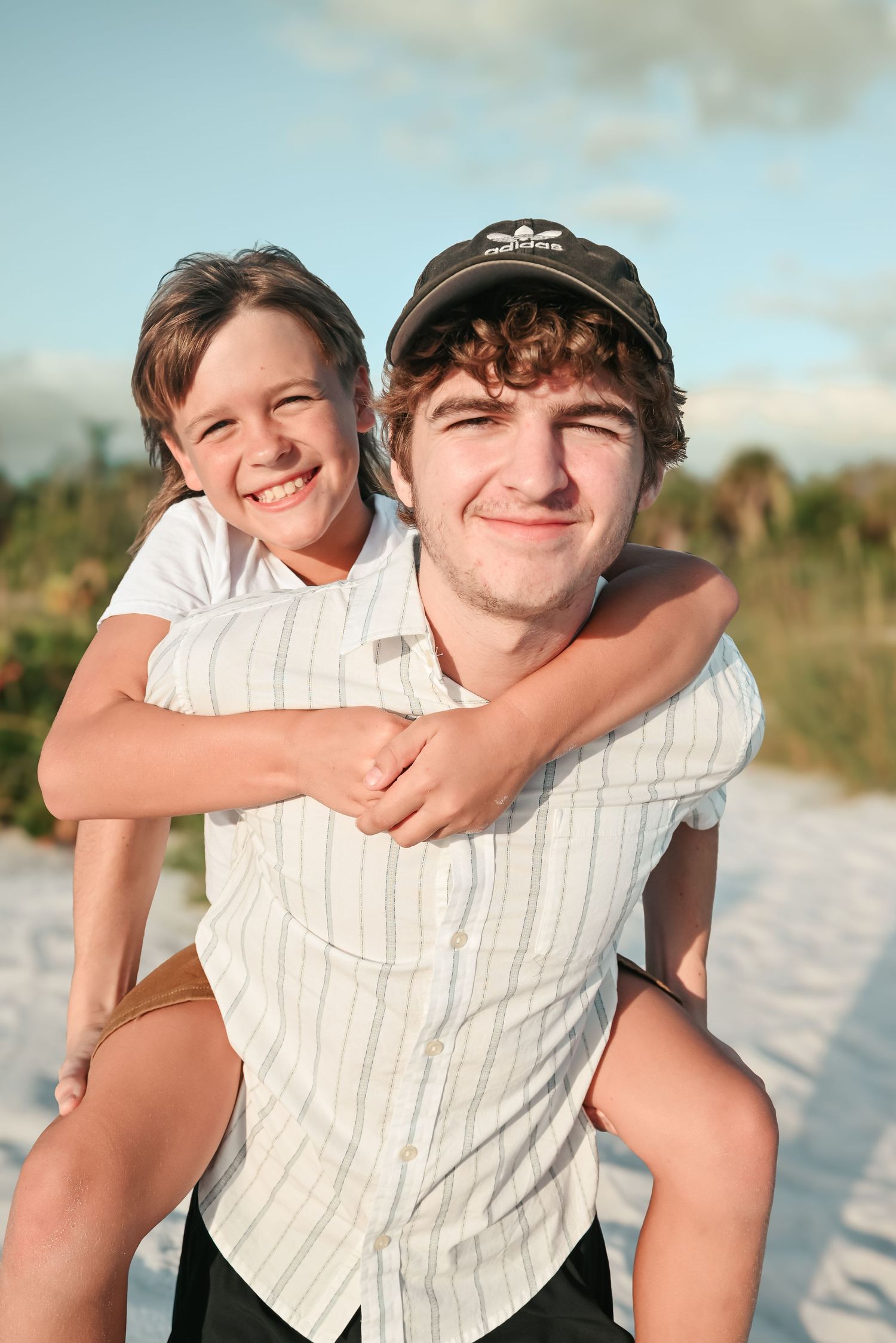 Two smiling brothers posing on a beach, younger boy giving a piggyback ride on his older brothers back.