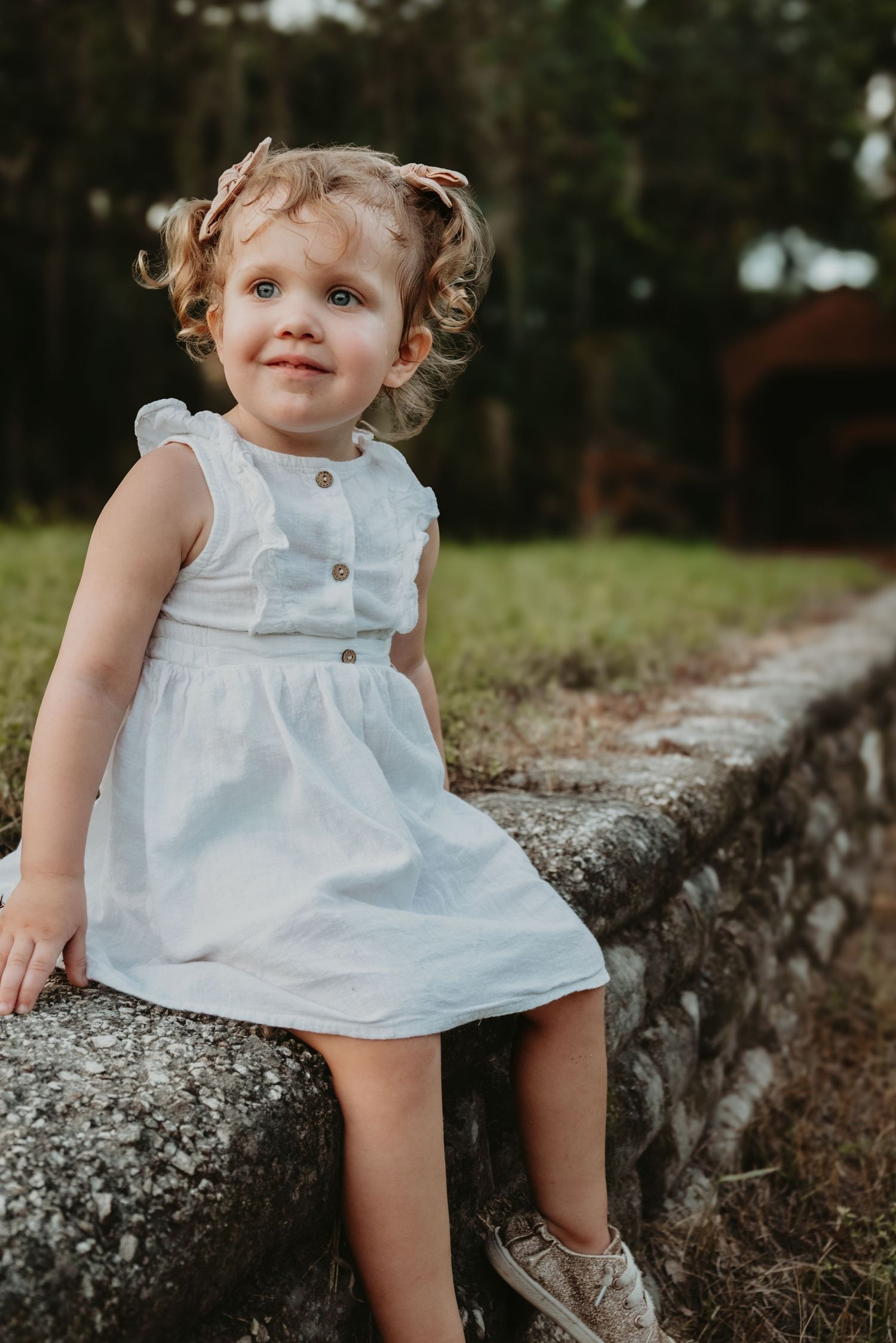 Adorable toddler girl in white dress sits on stone wall outdoors smiling at camera.