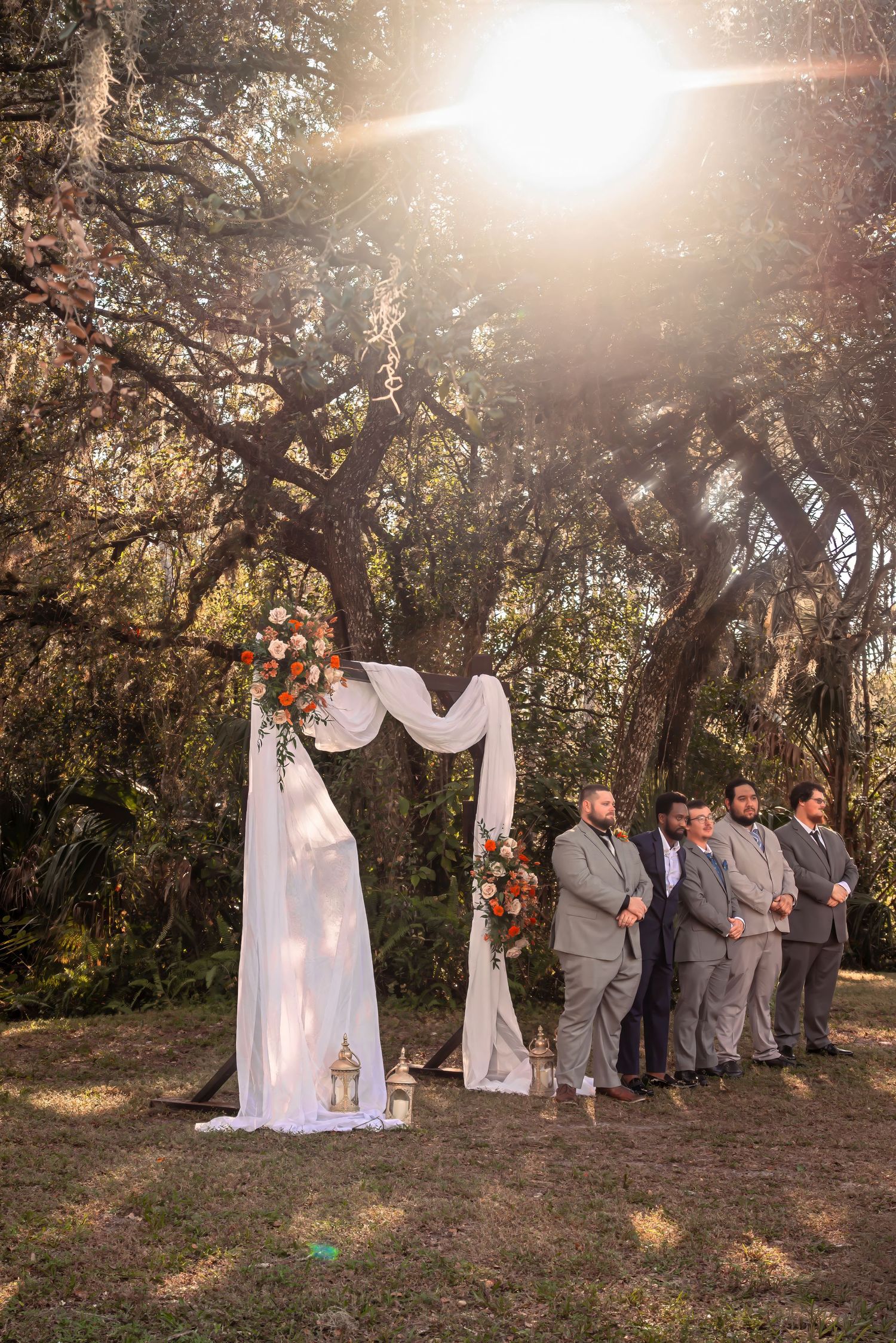 Elegant outdoor wedding arch adorned with orange and white florals as groomsmen await the ceremony.