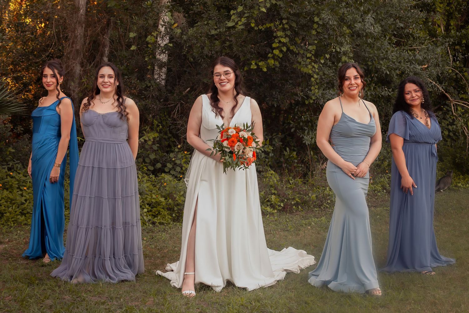 Bride in white gown holding orange bouquet with four bridesmaids in blue dresses outdoors.