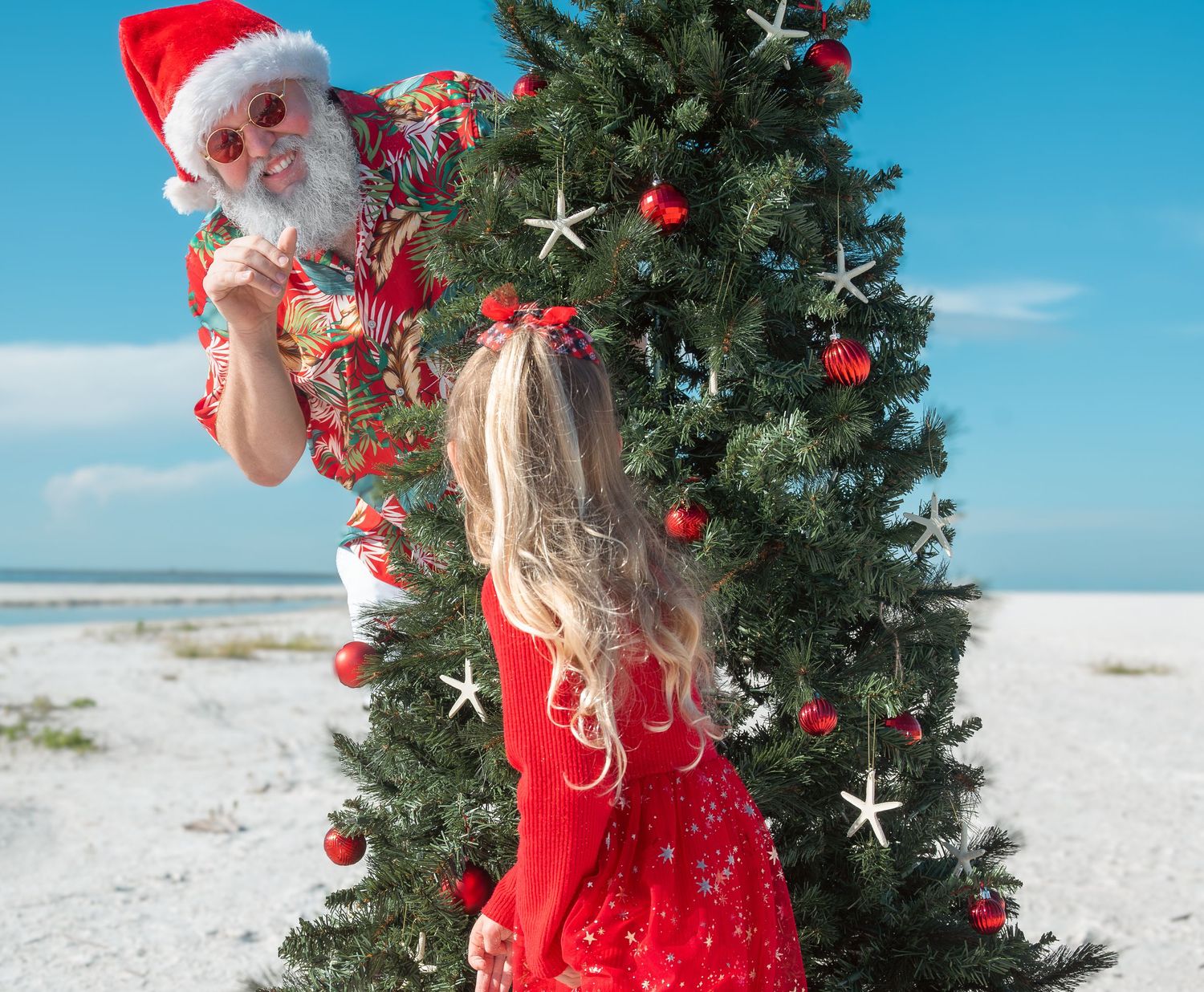 Little girl in red dress decorating a Christmas tree on a white sand beach.