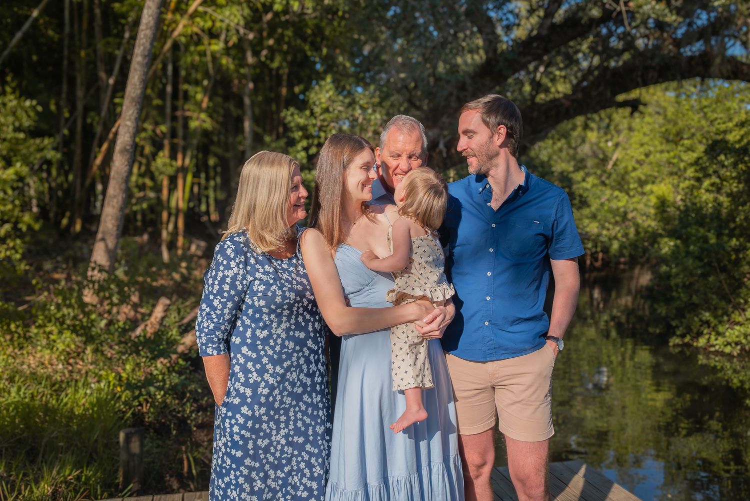 A happy family of five poses together near a pond in a lush outdoor setting.