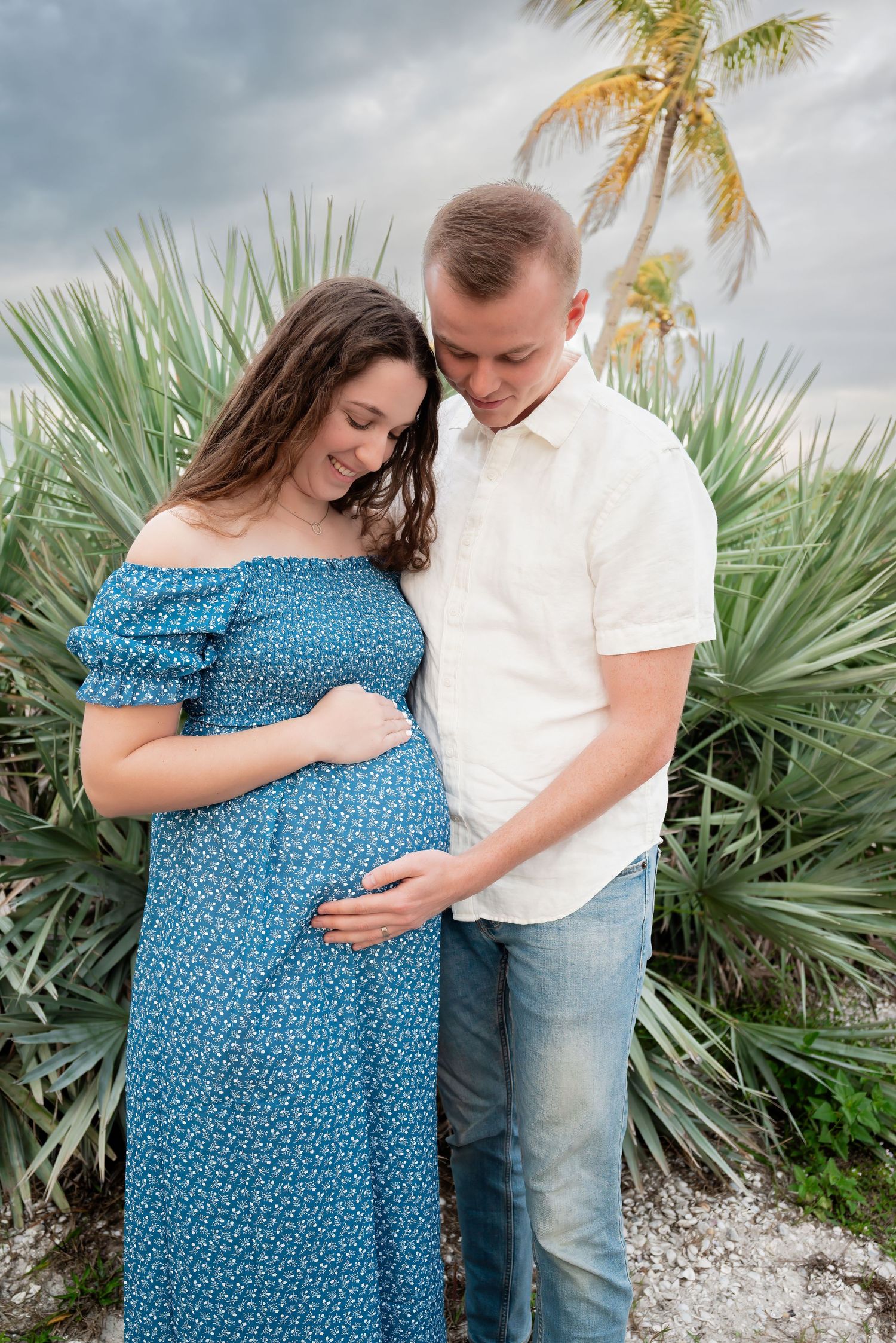 Pregnant couple embracing outdoors with tropical palm trees and cloudy sky background.