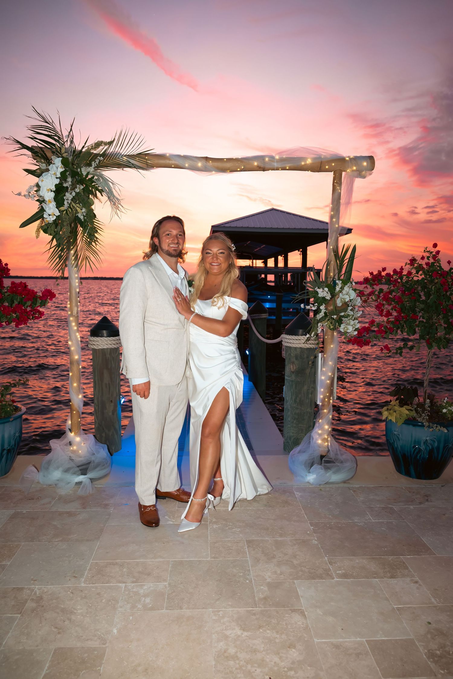 Couple poses under floral arch at waterfront sunset wedding ceremony in white dress and tan suit.