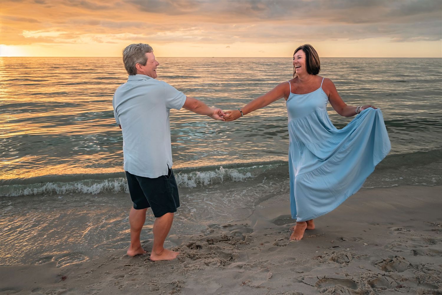 Romantic couple dancing and holding hands on a sandy beach during a golden sunset.