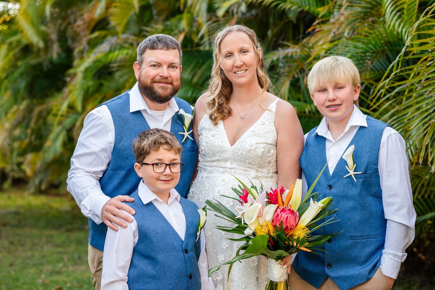 Wedding family portrait with bride, groom, and two boys in blue vests holding a tropical bouquet.