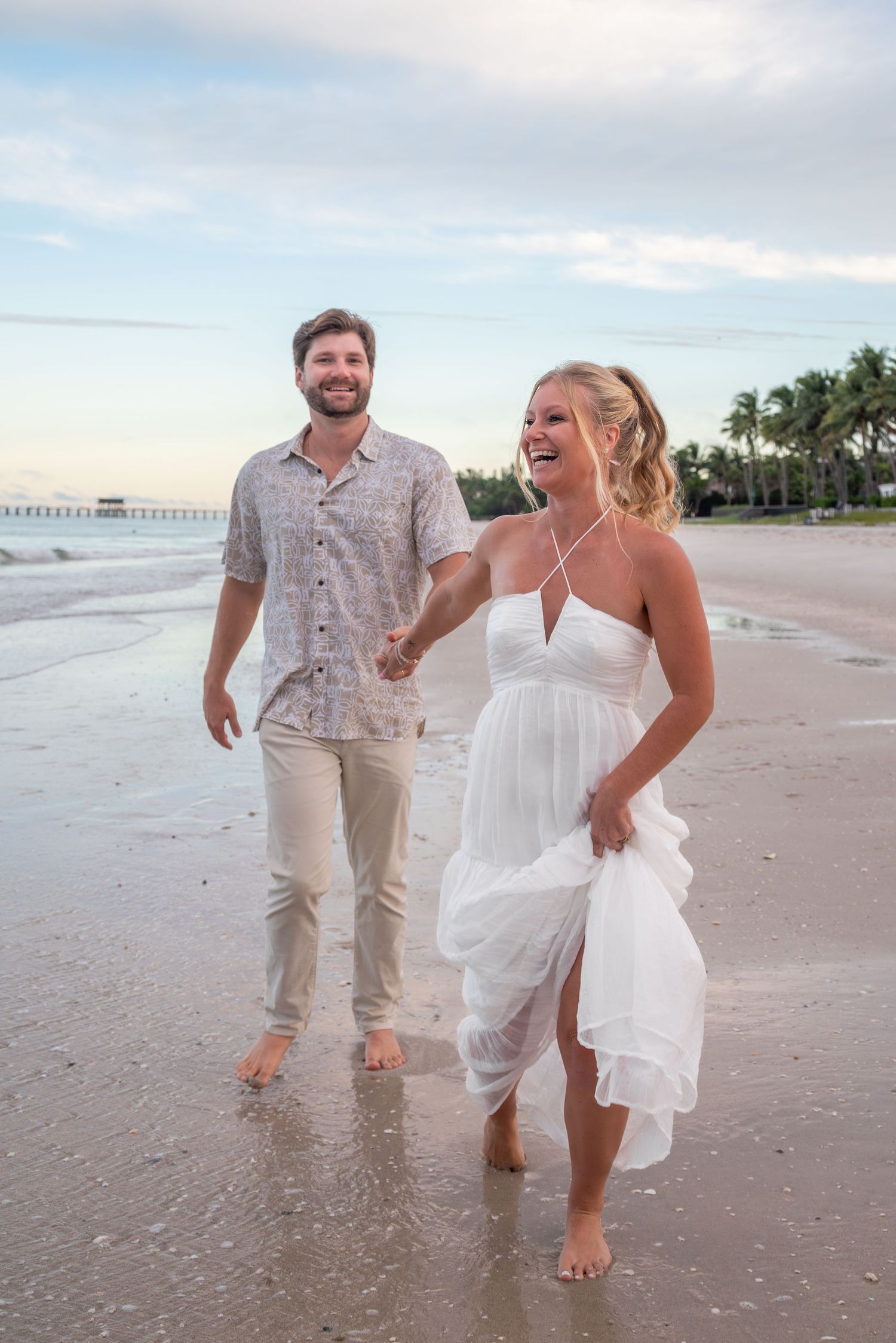 Happy couple walks barefoot on a tropical beach at sunset, woman in white dress laughing, man in casual shirt smiling.