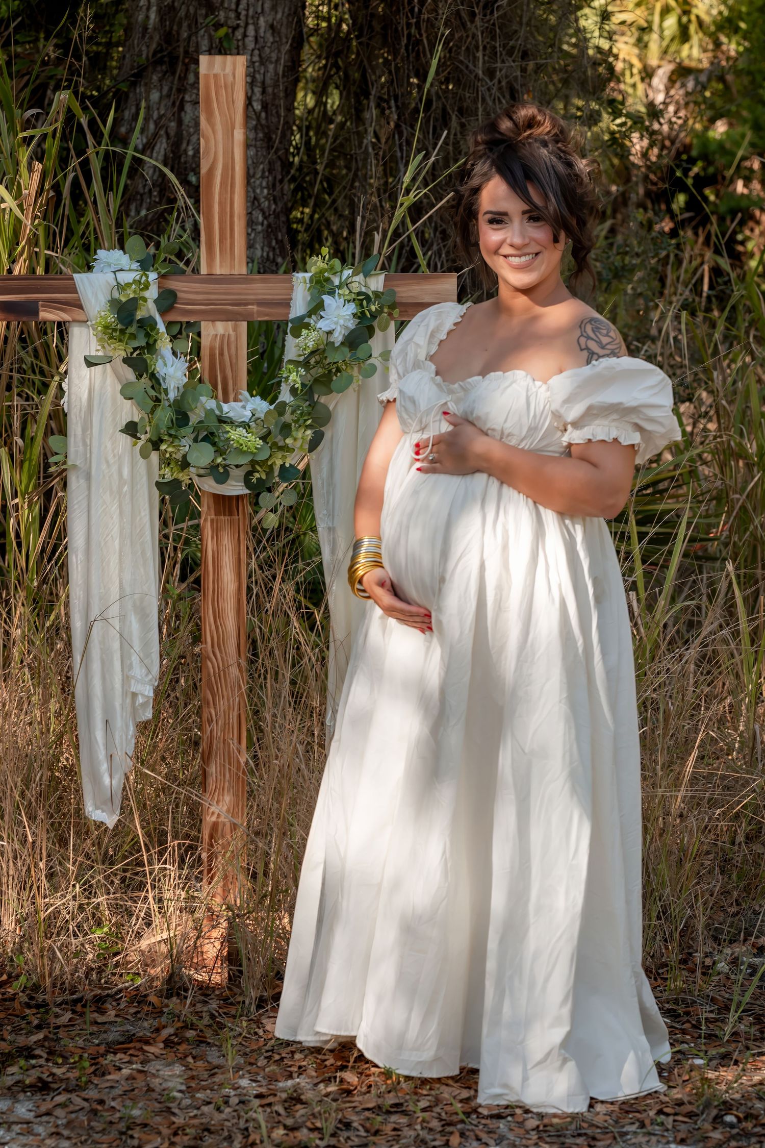 Smiling pregnant woman in white empire waist gown standing beside decorated wooden cross in nature.