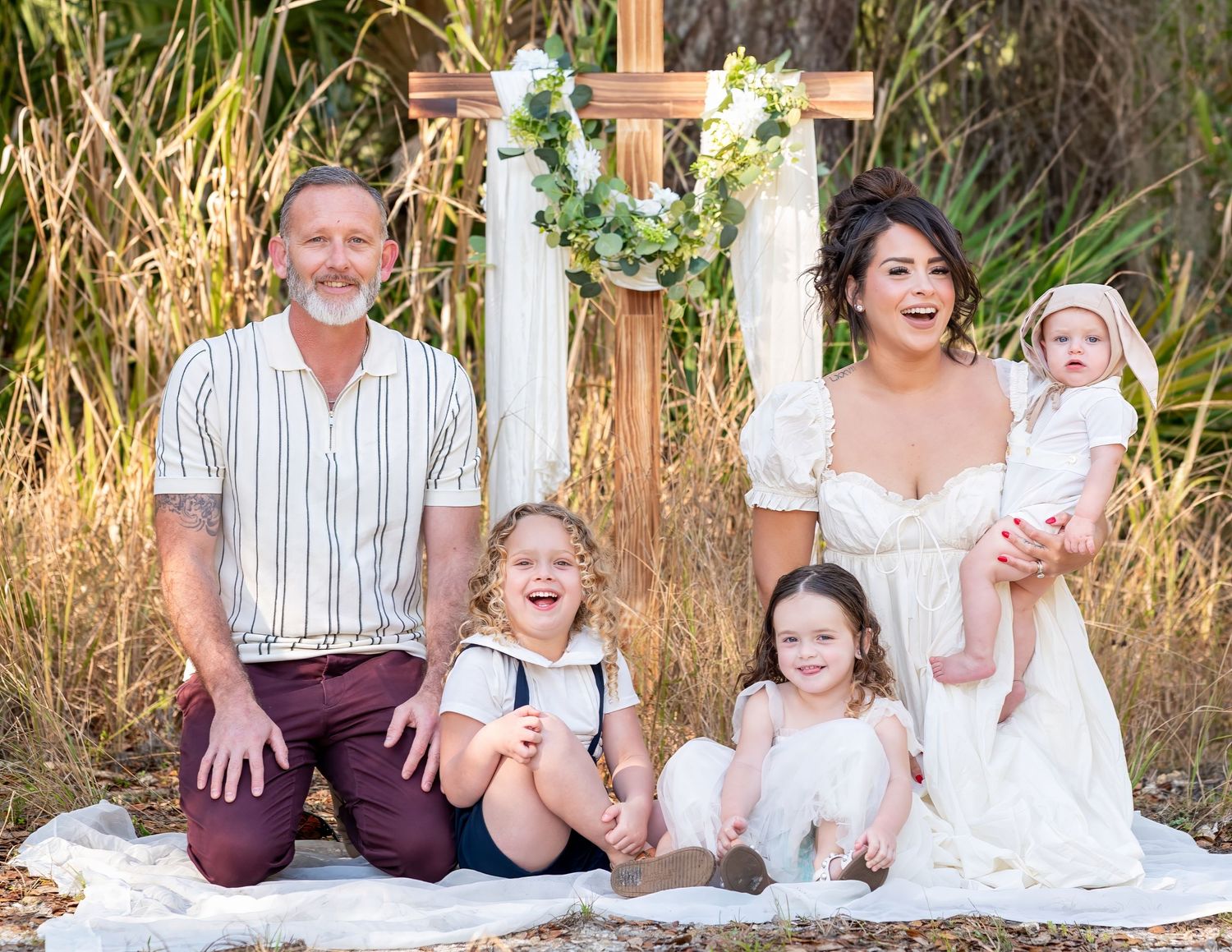 Smiling family of five with young children posing in front of a floral decorated wooden cross outdoors.