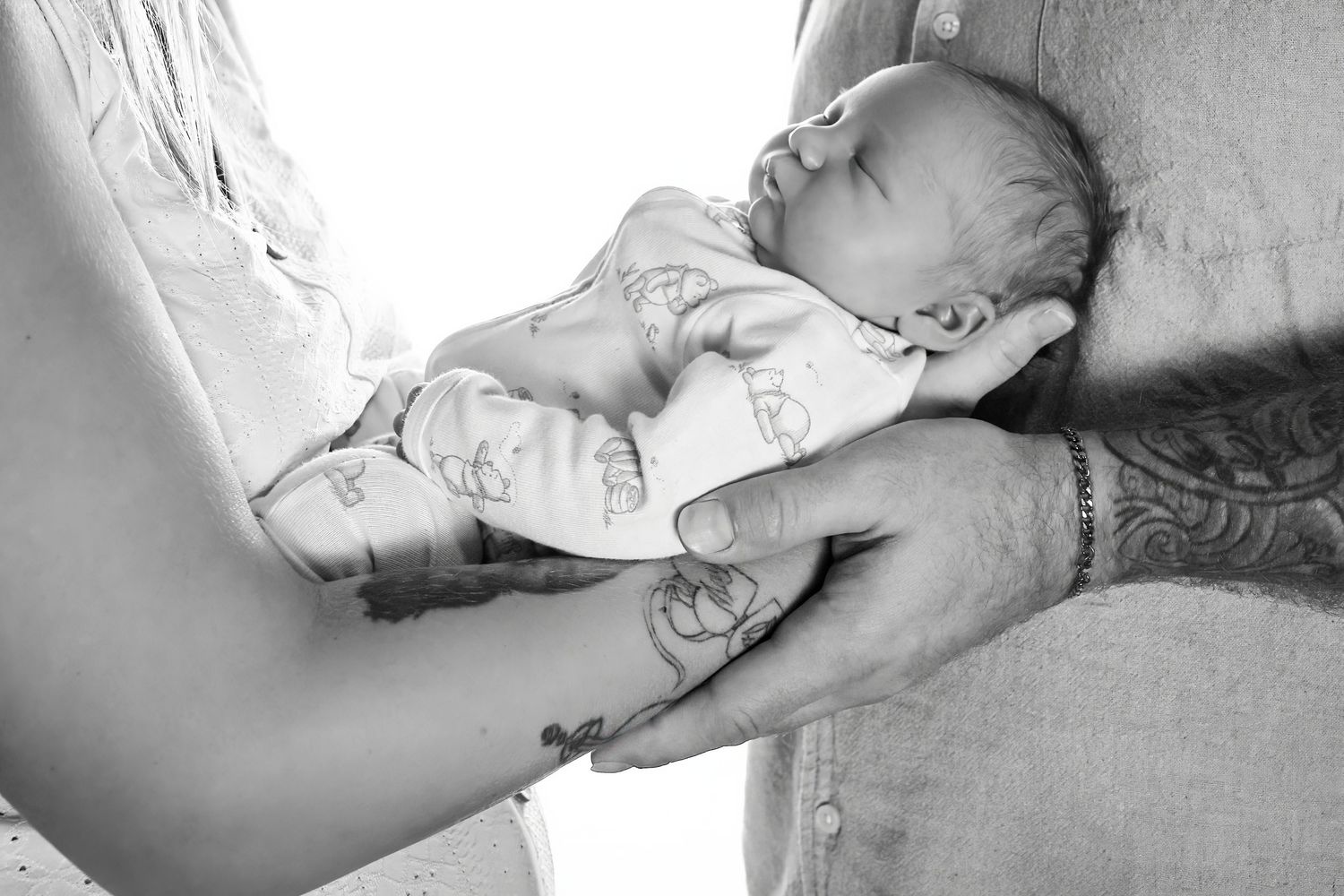 Black and white photo of a newborn baby cradled in parents hands, one arm showing a tattoo.