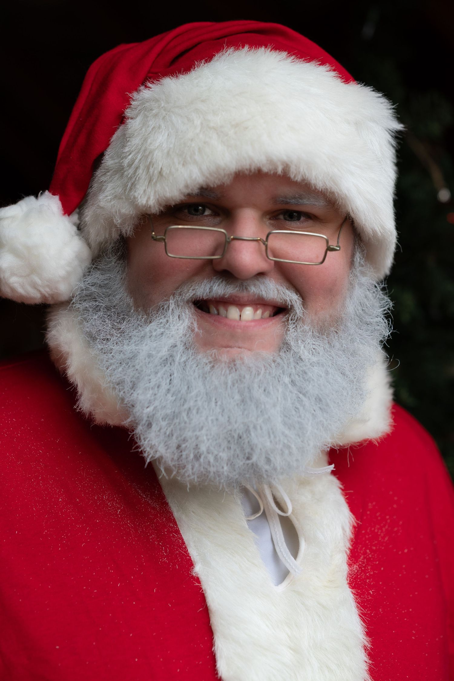 Smiling man dressed as Santa Claus wearing red suit, white beard, glasses, and classic red hat with white trim.