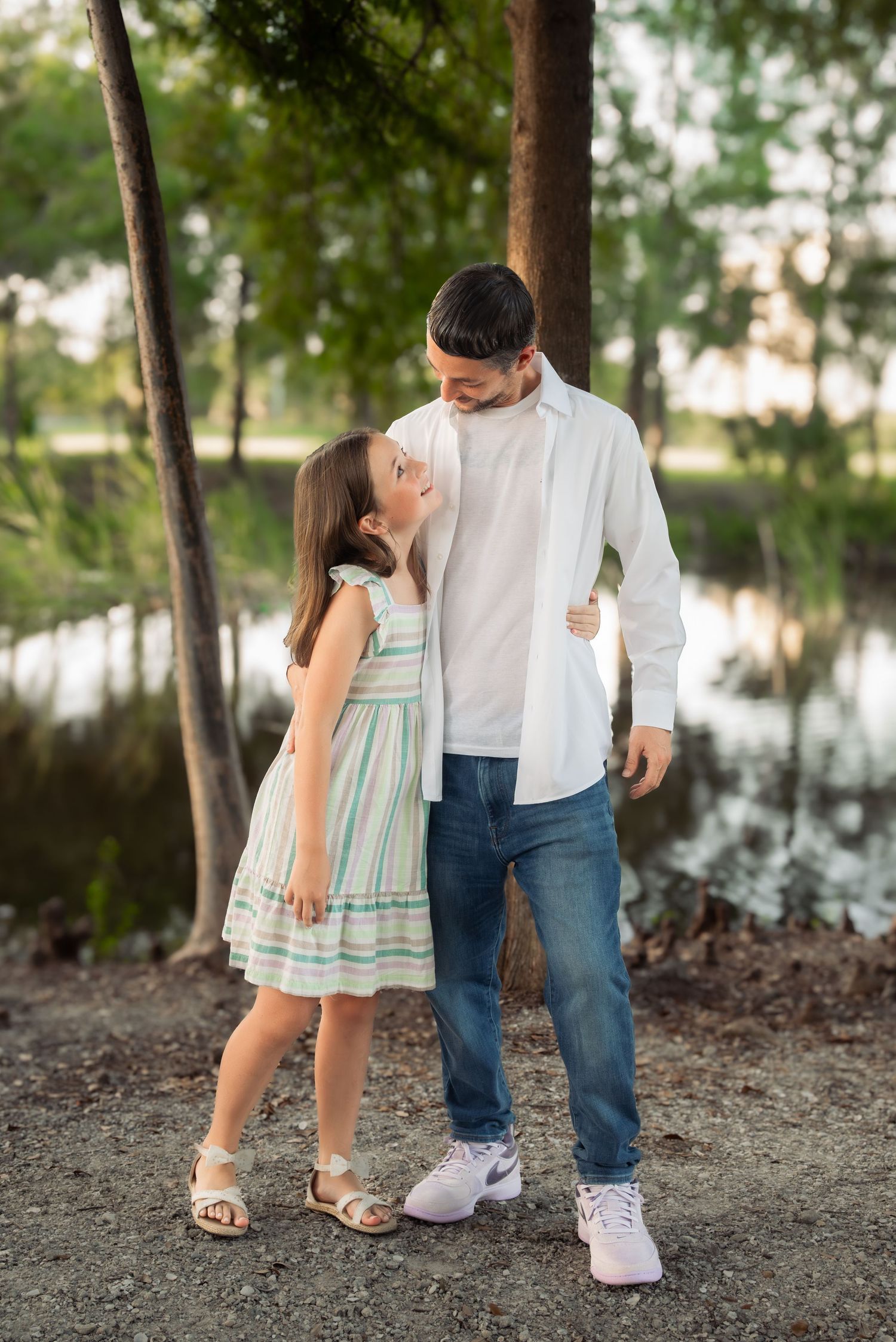 Sweet young girl looking up at her father while standing together near a peaceful lakeside park setting.