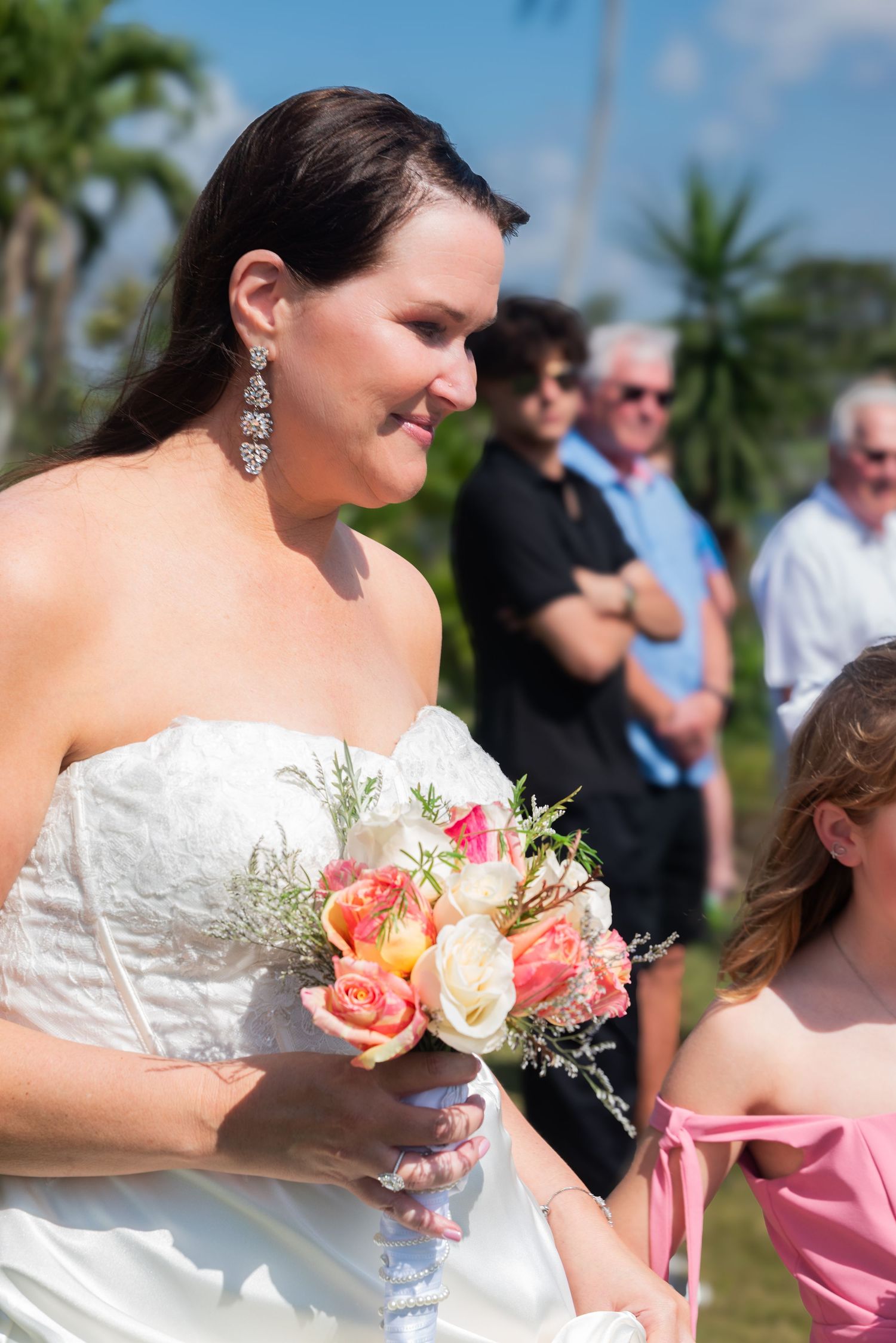 Bride in white strapless gown holds colorful rose bouquet while smiling during outdoor wedding ceremony.