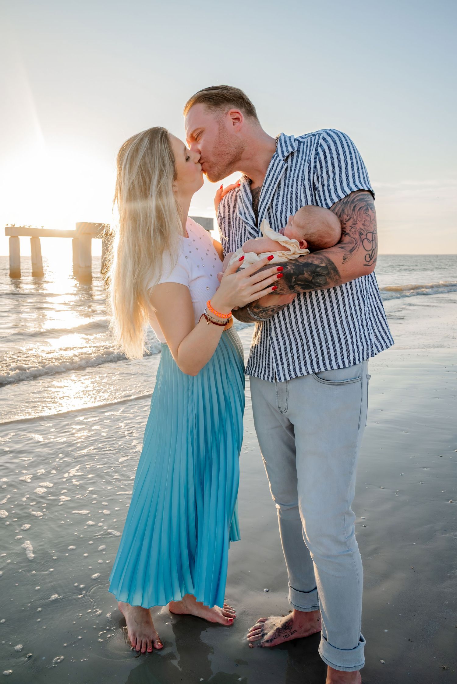 Couple kissing on beach at sunset while holding newborn baby near ocean pier.