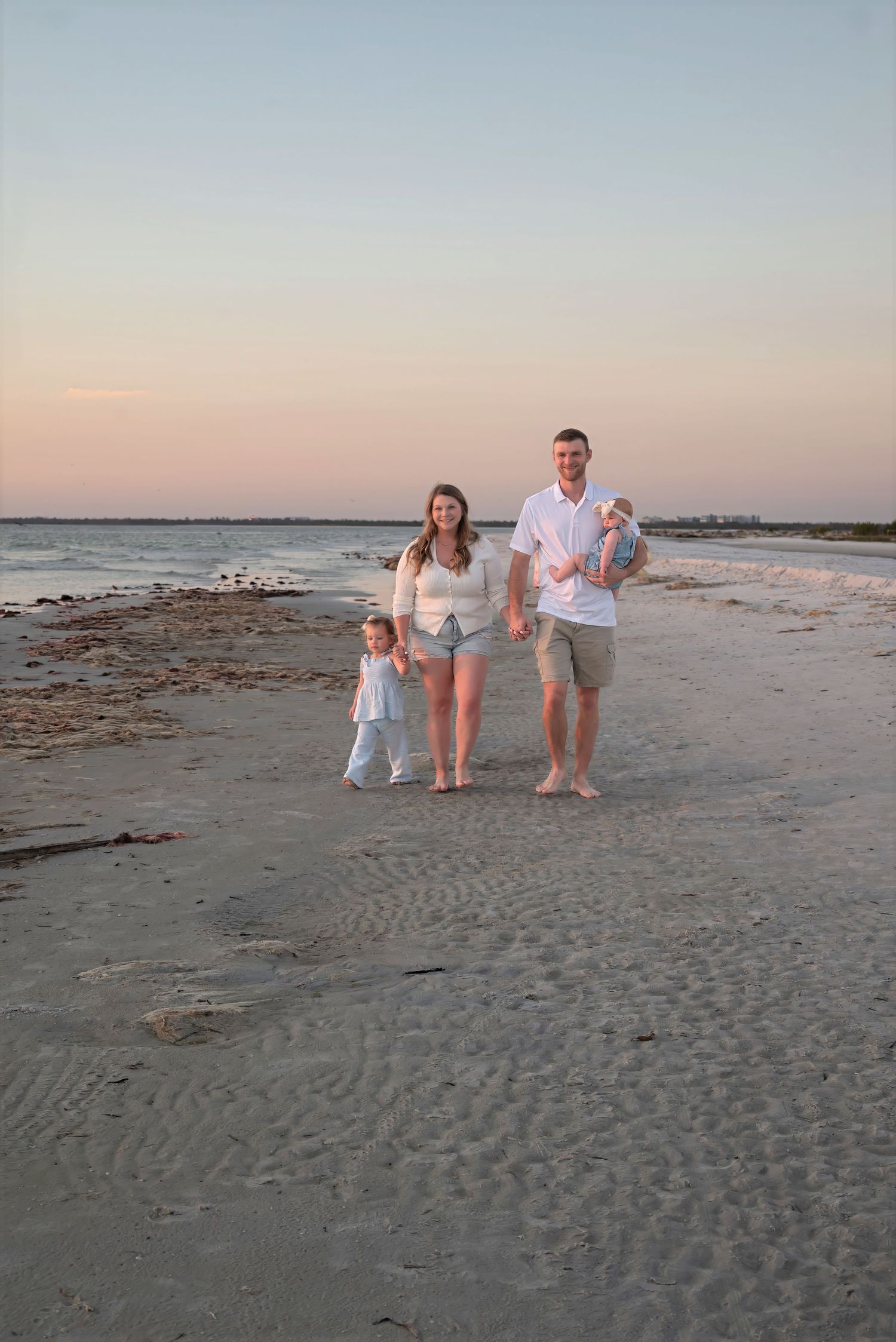 Young family with toddler and baby posing on sandy beach at sunset.