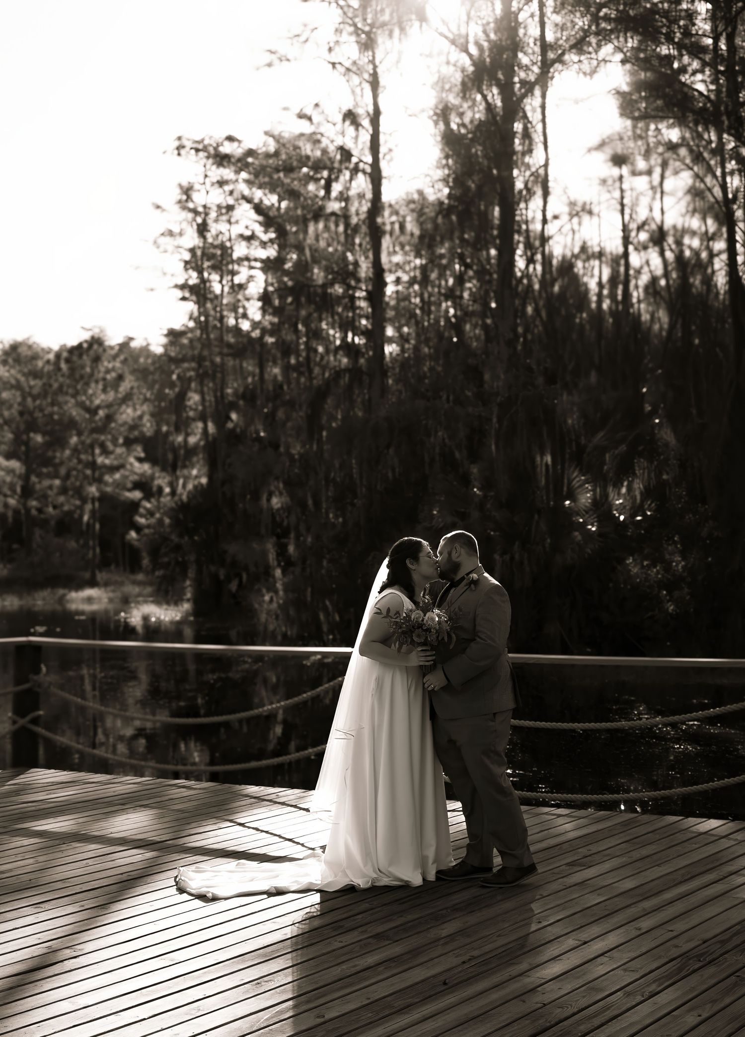 Black and white romantic wedding kiss photo on a wooden deck surrounded by towering trees and natural scenery.
