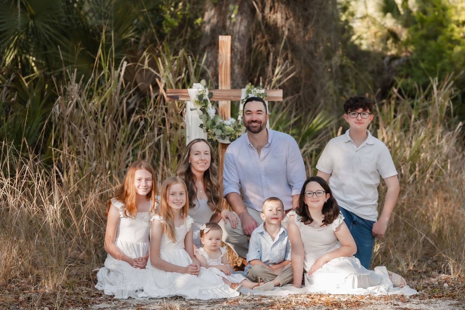 Large family of eight posing outdoors near a wooden cross decorated with flowers in a natural setting.