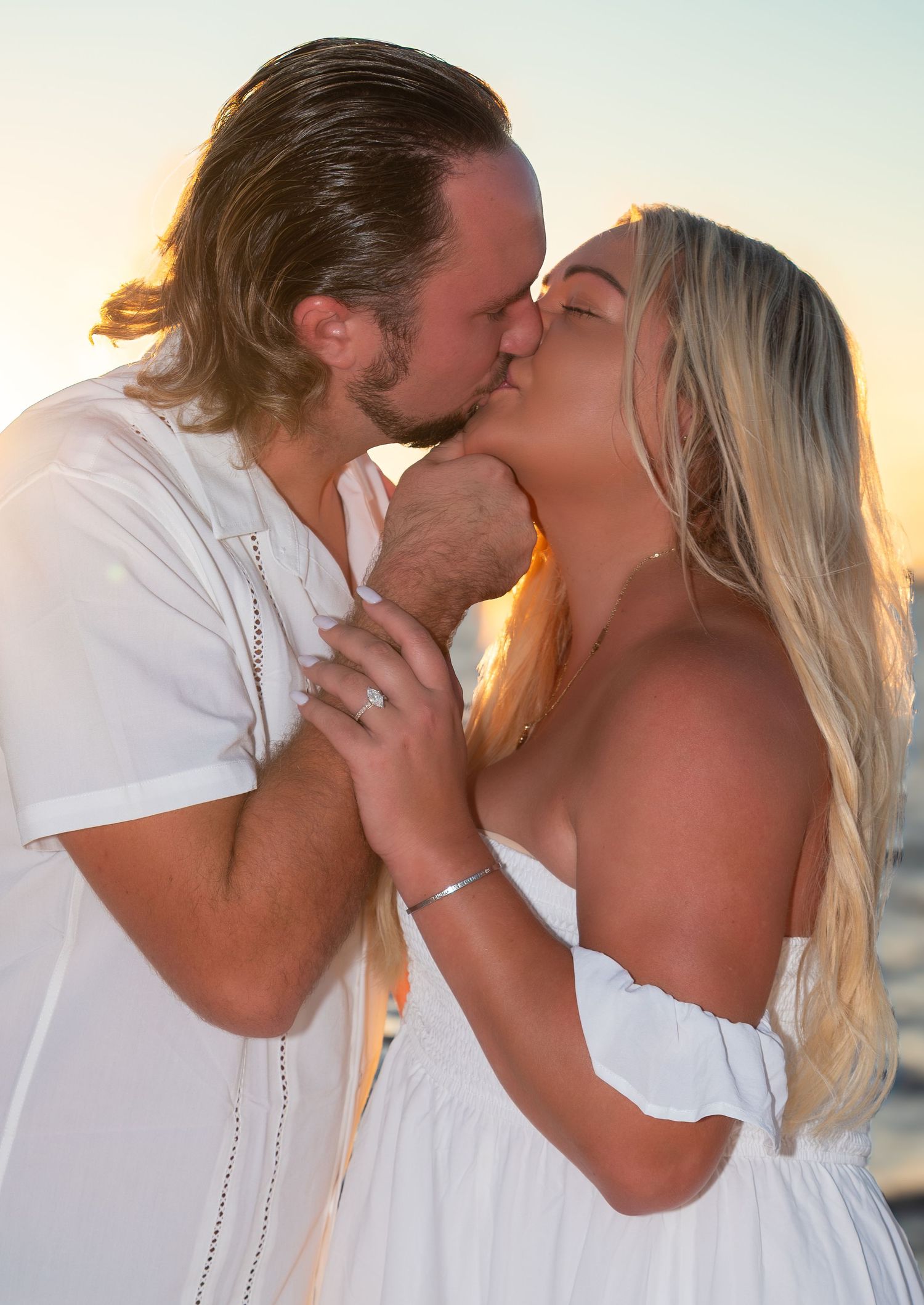 Engaged couple sharing a romantic kiss at sunset, woman displaying her engagement ring.