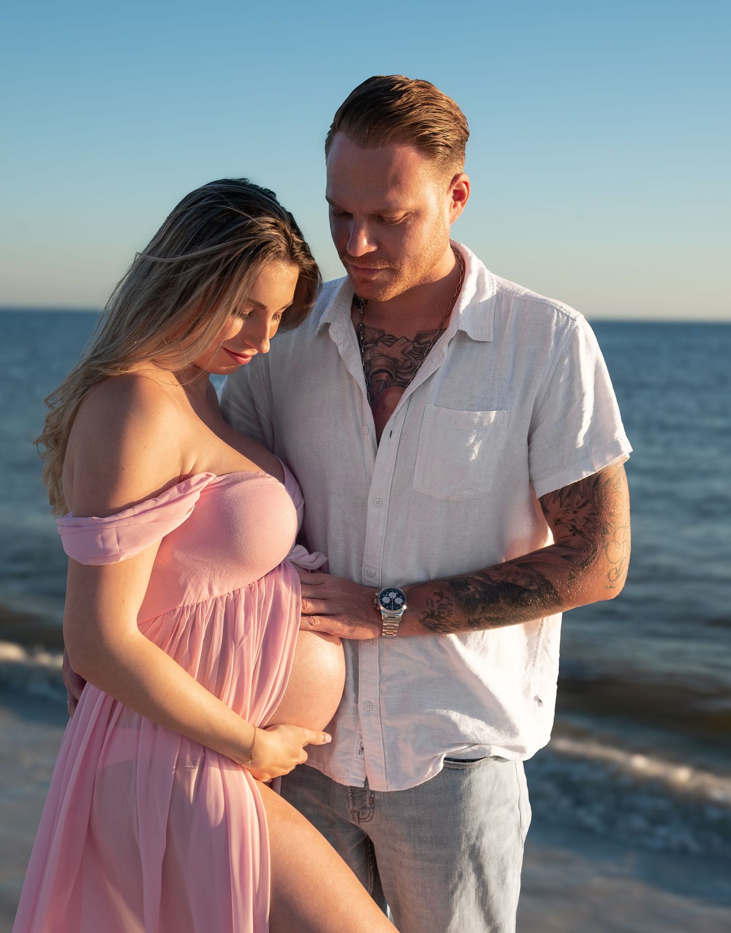 Tattooed man and pregnant woman in pink dress sharing a tender moment on the beach.