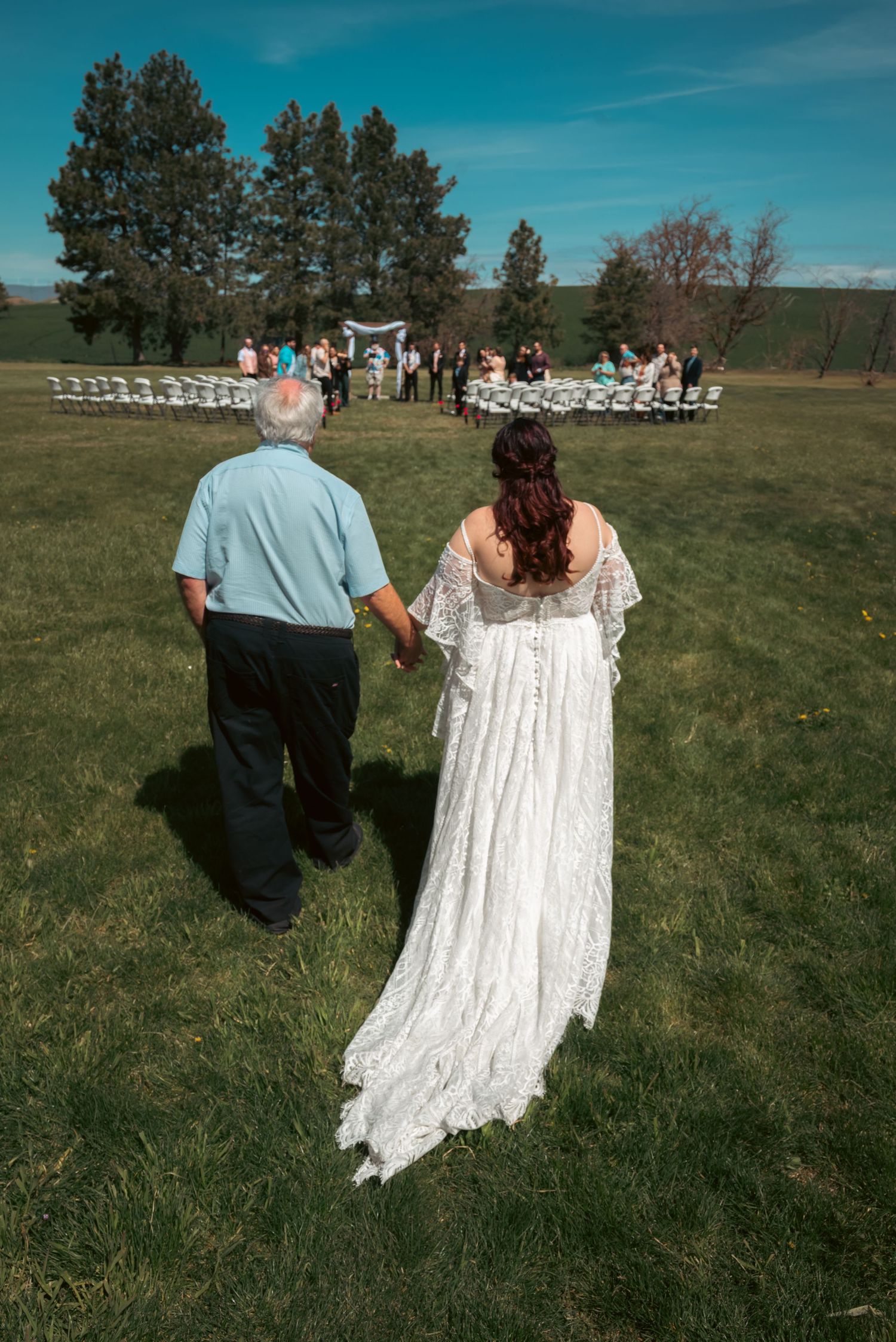 Bride in lace gown walks hand-in-hand with older man toward outdoor wedding ceremony on green lawn.