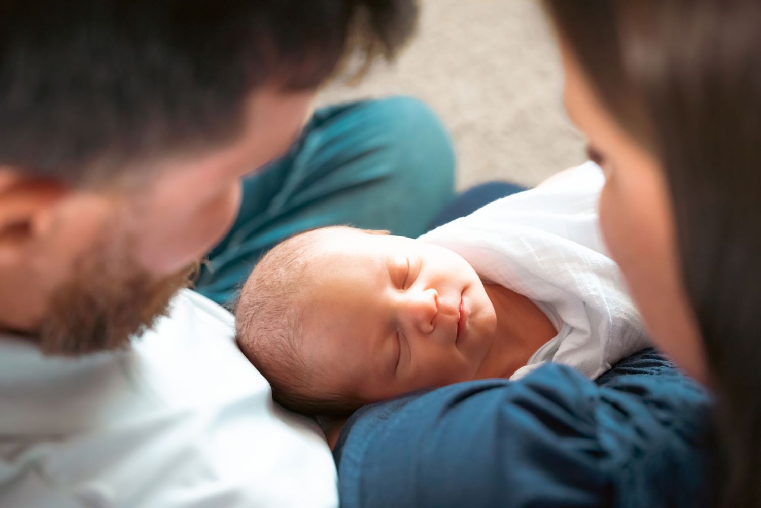 Parents lovingly gazing at their sleeping newborn baby wrapped in a white blanket.
