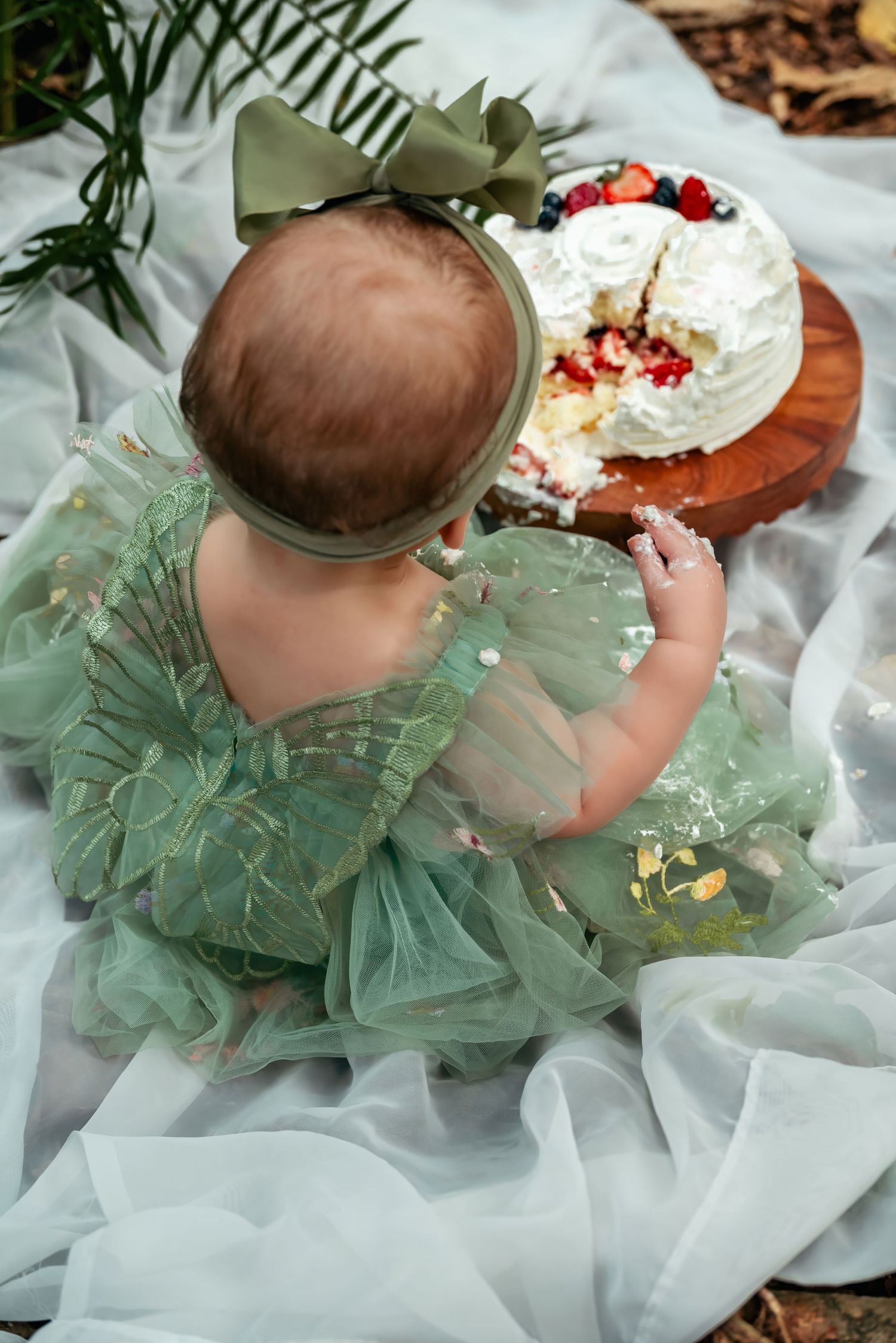 Baby girl in green tulle dress with bow headband reaching for a white cake with berries on a wooden stand outdoors.