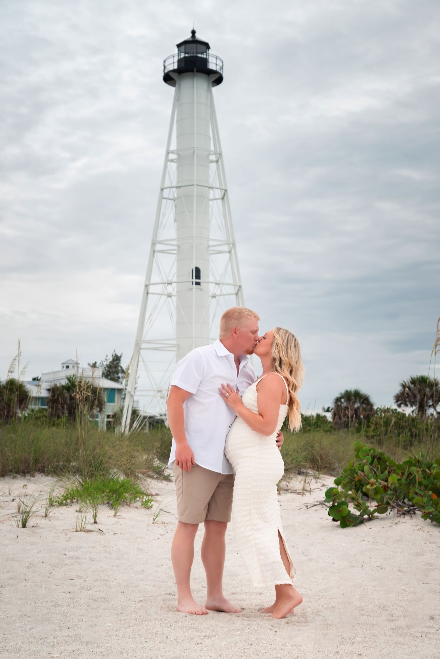Pregnant couple kissing barefoot on sandy beach with white lighthouse backdrop under cloudy sky.