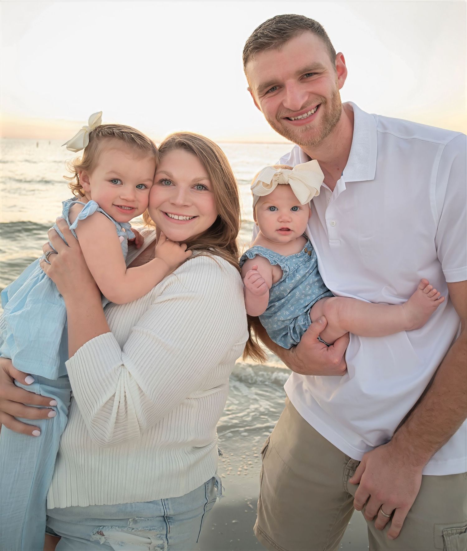 Young family of four smiles together on a beach at sunset, parents holding two small daughters.