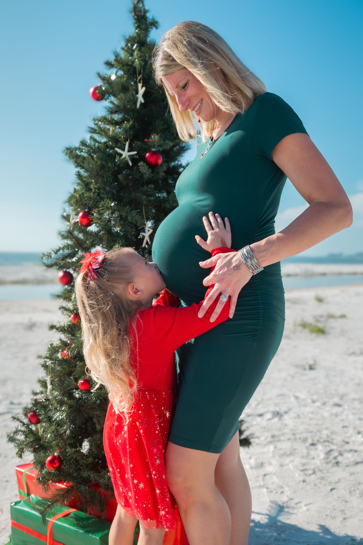 Young girl hugs pregnant mothers baby bump beside decorated Christmas tree on sunny beach in matching red outfits.