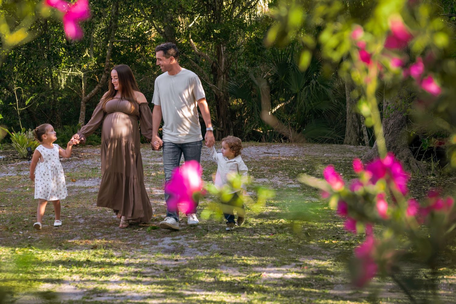 Happy expecting family with two young children walks hand in hand on a sunlit path framed by pink flowers.