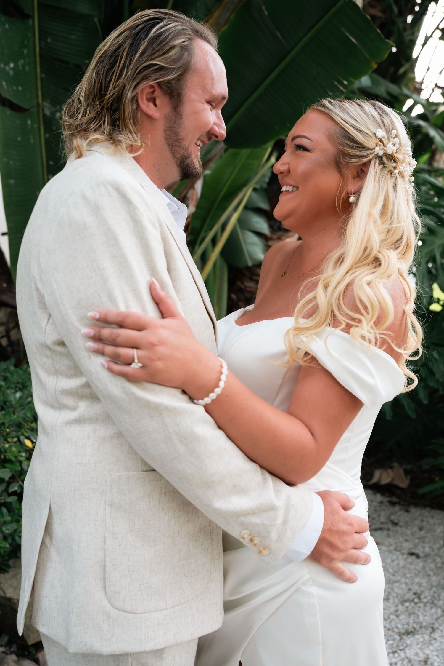 A smiling bride and groom in cream wedding attire embrace outdoors with tropical greenery in the background.