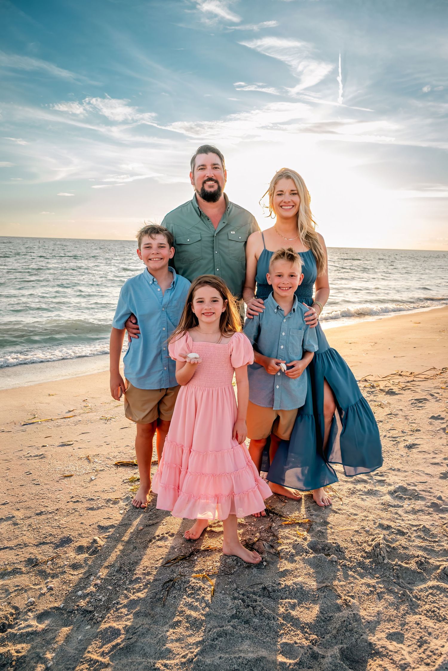 Family of five posing together on a sandy beach at sunset, wearing coordinated blue and pink outfits.