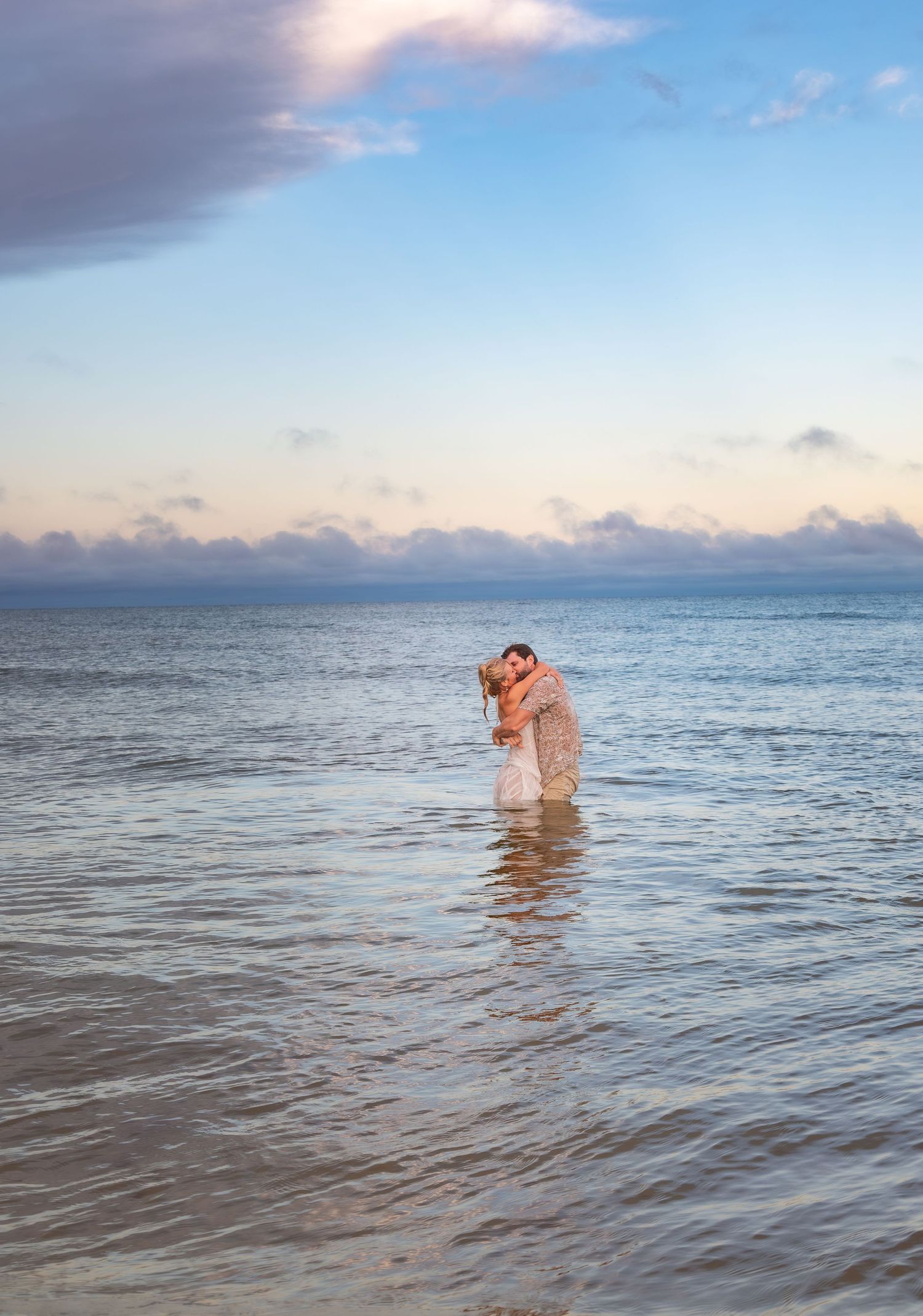 Couple embracing and kissing while standing in shallow ocean water at sunset with a pastel sky backdrop.